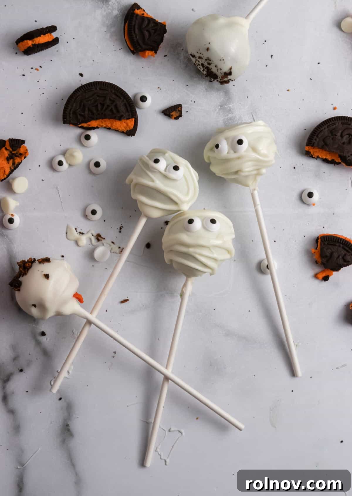A charming display of Halloween cake pops alongside ingredients like crushed Oreos, white chocolate chips, and candy eyeballs on a rustic kitchen counter.
