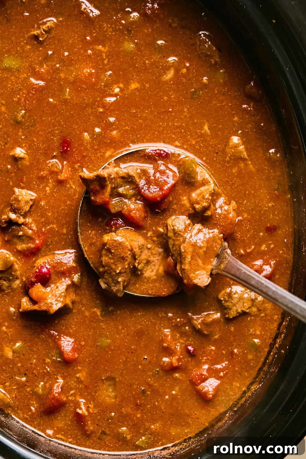 A ladle scooping a rich portion of tender beef brisket chili from a slow cooker, highlighting the hearty texture and ingredients.