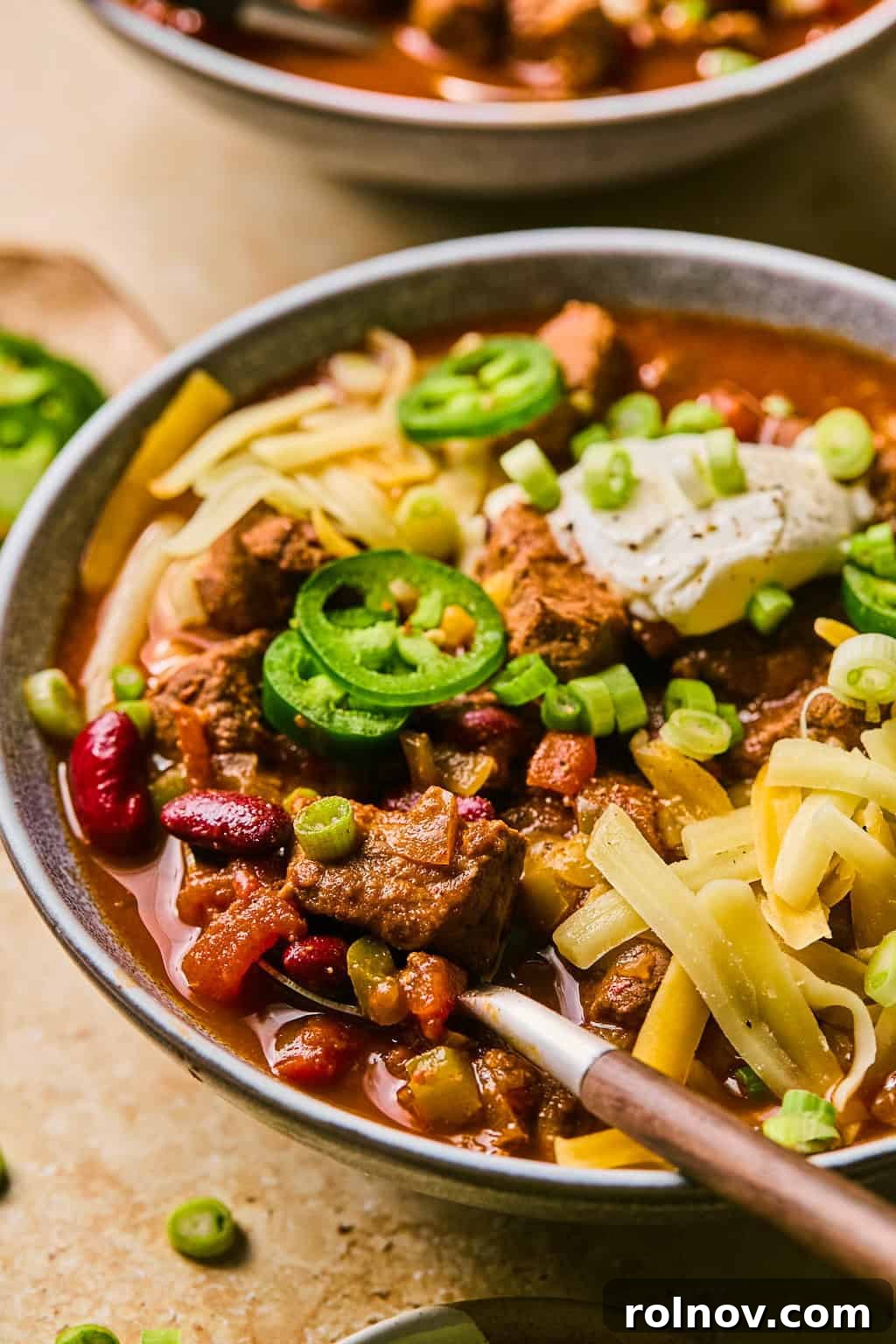 A close-up of a bowl of slow cooker beef brisket chili, topped with vibrant orange cheese and a spoon ready to scoop, showcasing its hearty texture.