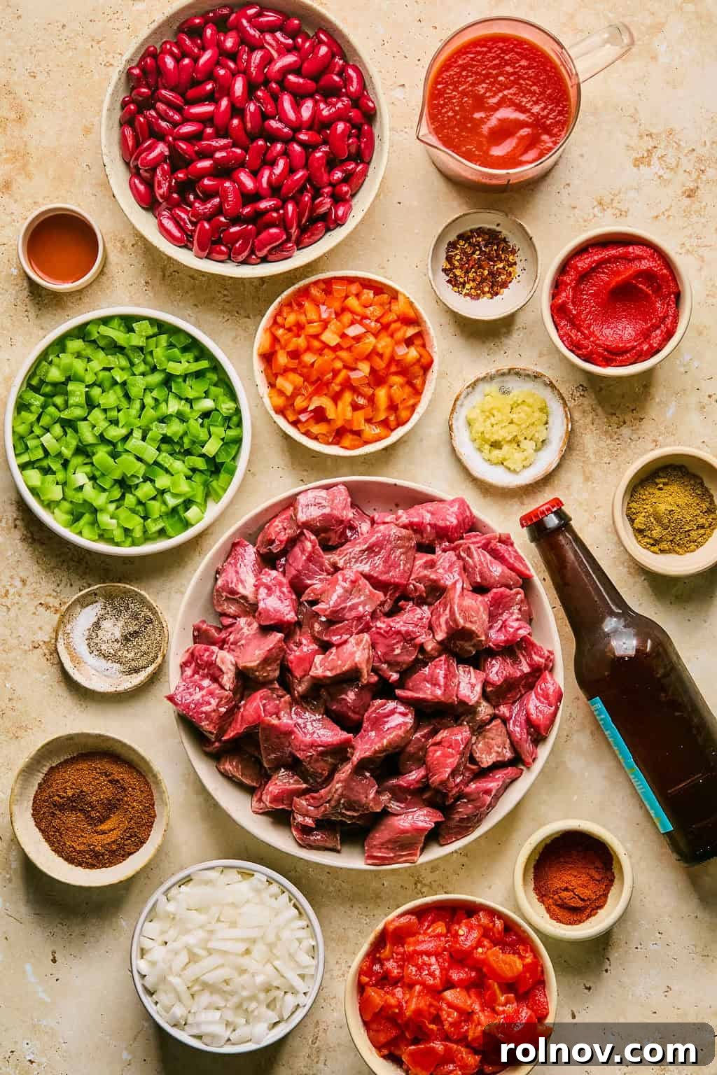 A vibrant flat lay of ingredients for slow cooker beef brisket chili, including cubed beef brisket, kidney beans, diced tomatoes, colorful bell peppers, a can of IPA beer, garlic cloves, and various spices.