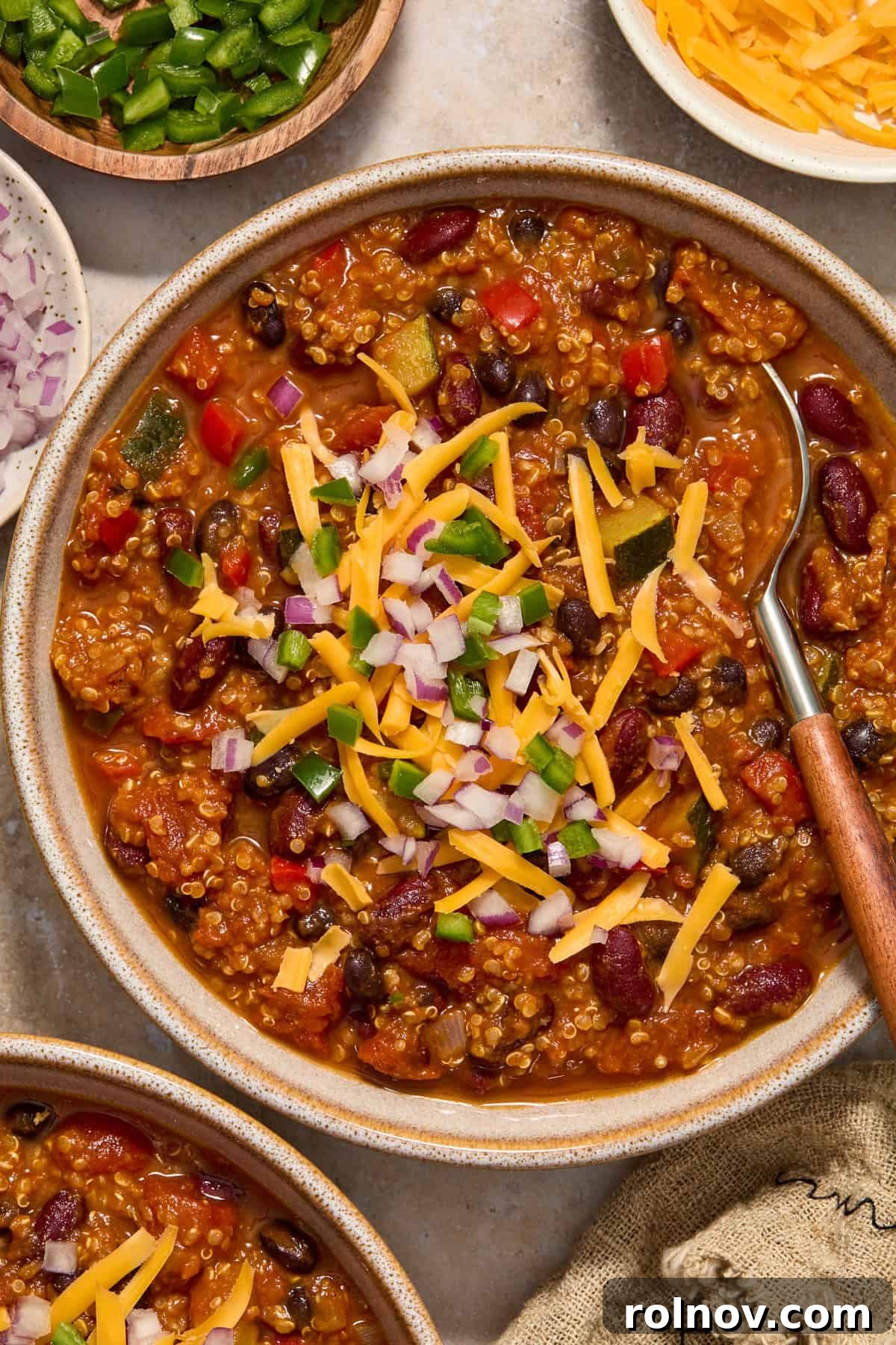 A bowl of pumpkin chili recipe generously topped with shredded cheese, chopped red onion, and sliced jalapeño.
