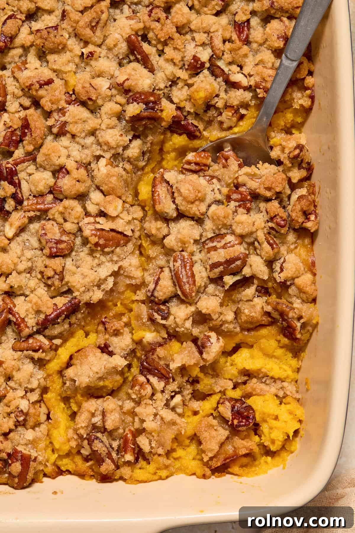 A serving spoon expertly scoops into the creamy, pecan crumble-topped butternut squash mixture in a rustic baking pan, highlighting its appealing texture.
