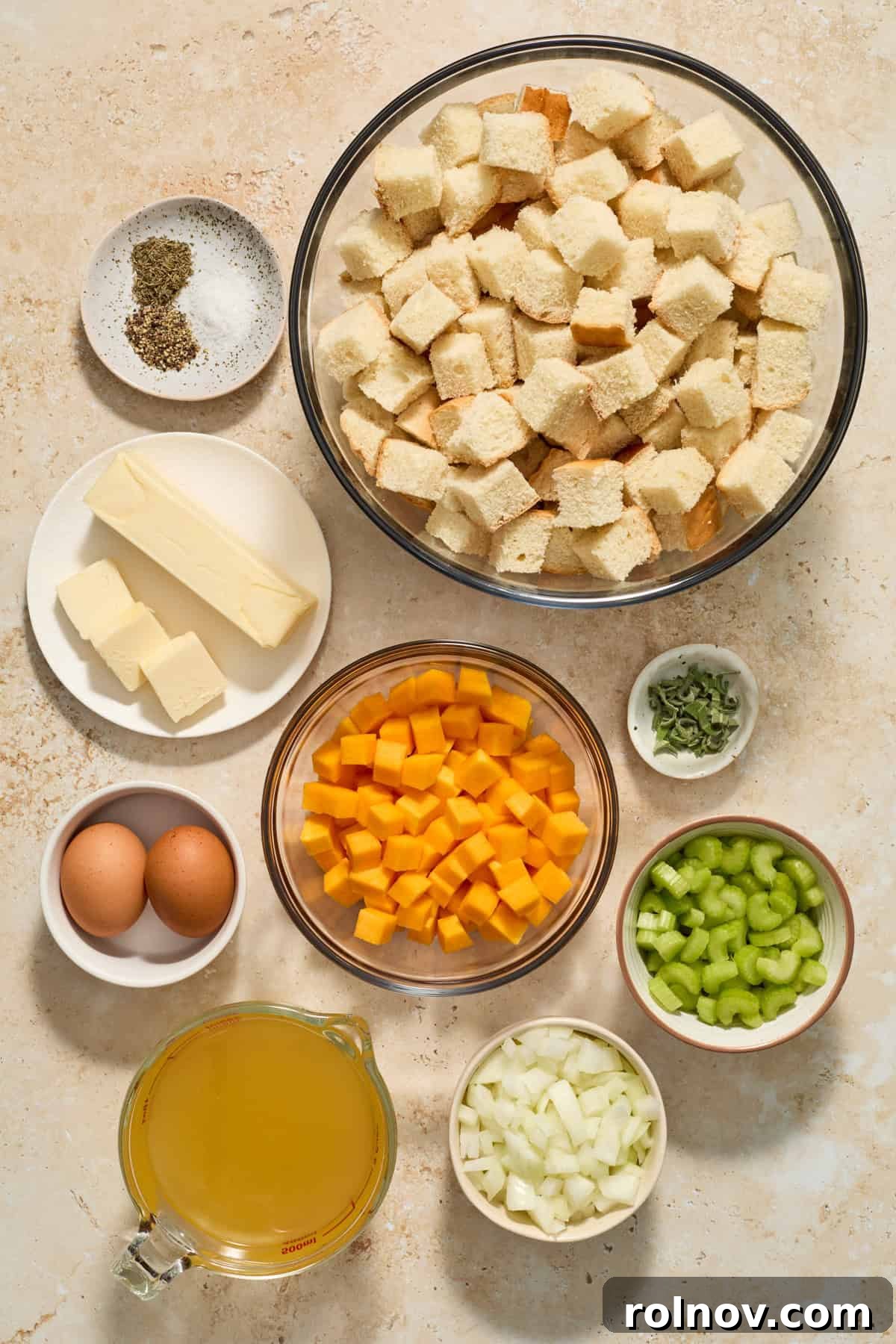 Assorted stuffing ingredients laid out on a counter, including cubed bread, chopped celery, onions, fresh herbs, and a bowl of broth and butter.