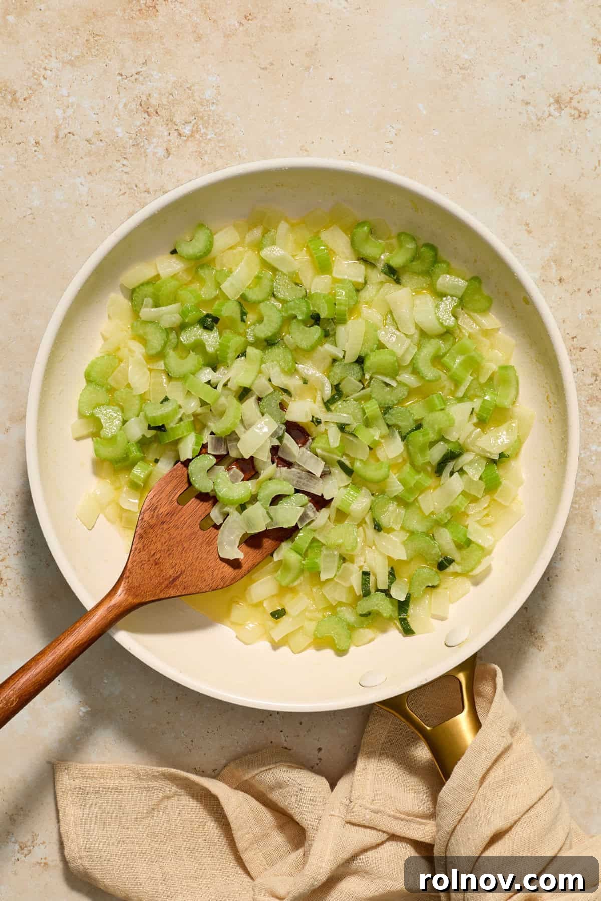 Sautéed onion and celery simmering in melted butter with fresh herbs in a skillet, stirred with a wooden spatula.