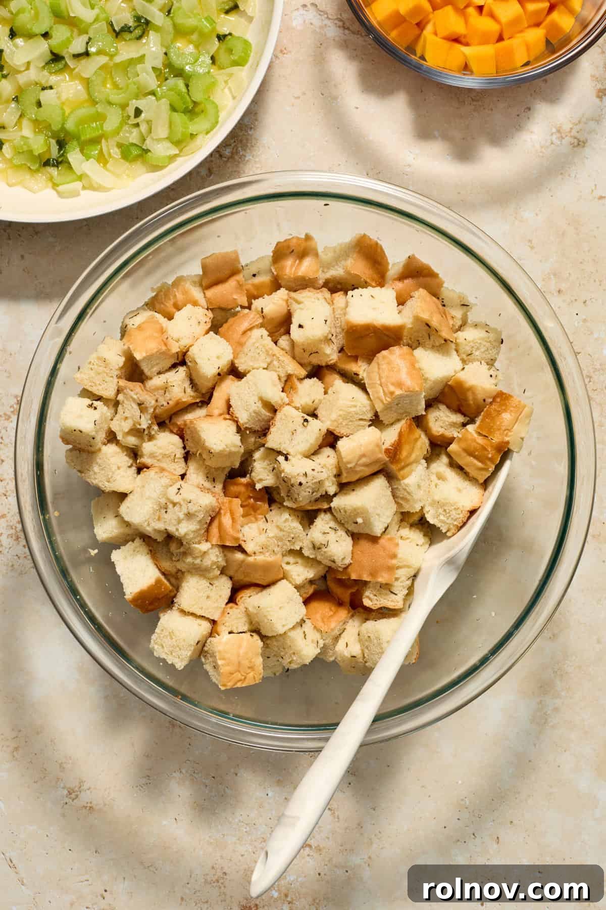 Cubed bread in a large glass bowl with a spatula, next to a skillet of cooked vegetables and a separate bowl of butternut squash cubes.