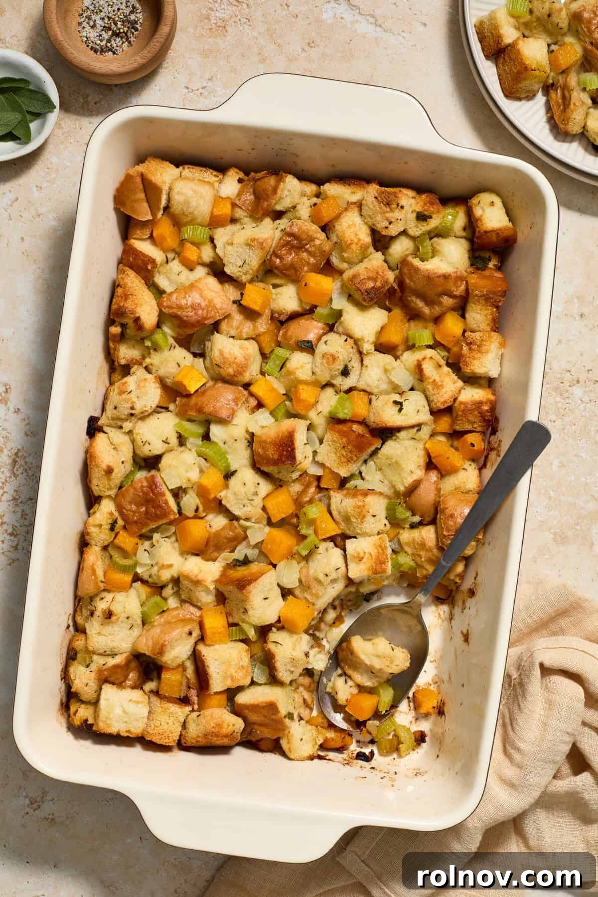A baking pan of freshly baked butternut squash stuffing with a large serving spoon scooping out a portion, highlighting its golden crust and tender interior.