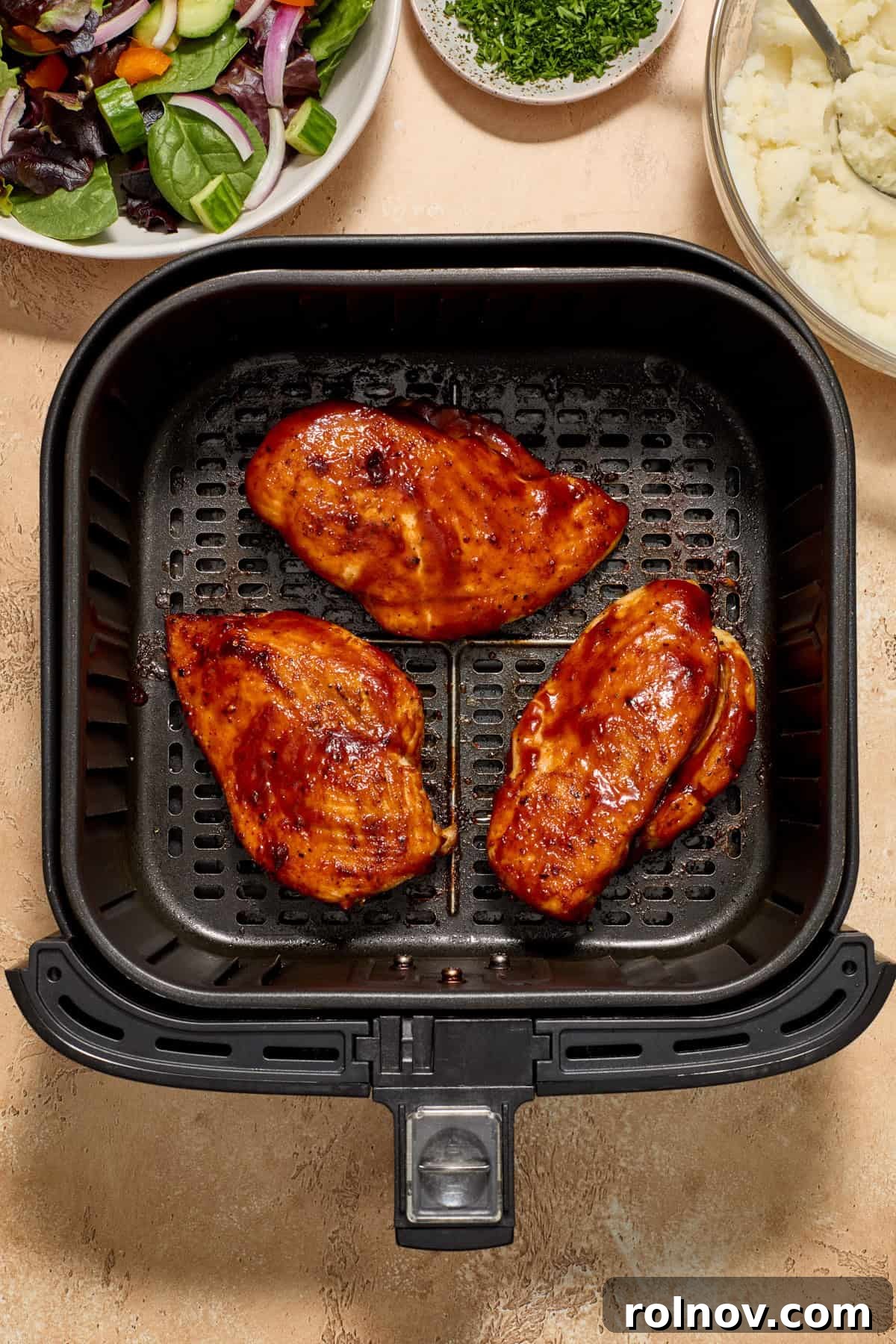Cooked air fryer bbq chicken breast in air fryer basket with potatoes and salad in bowls beside the air fryer.