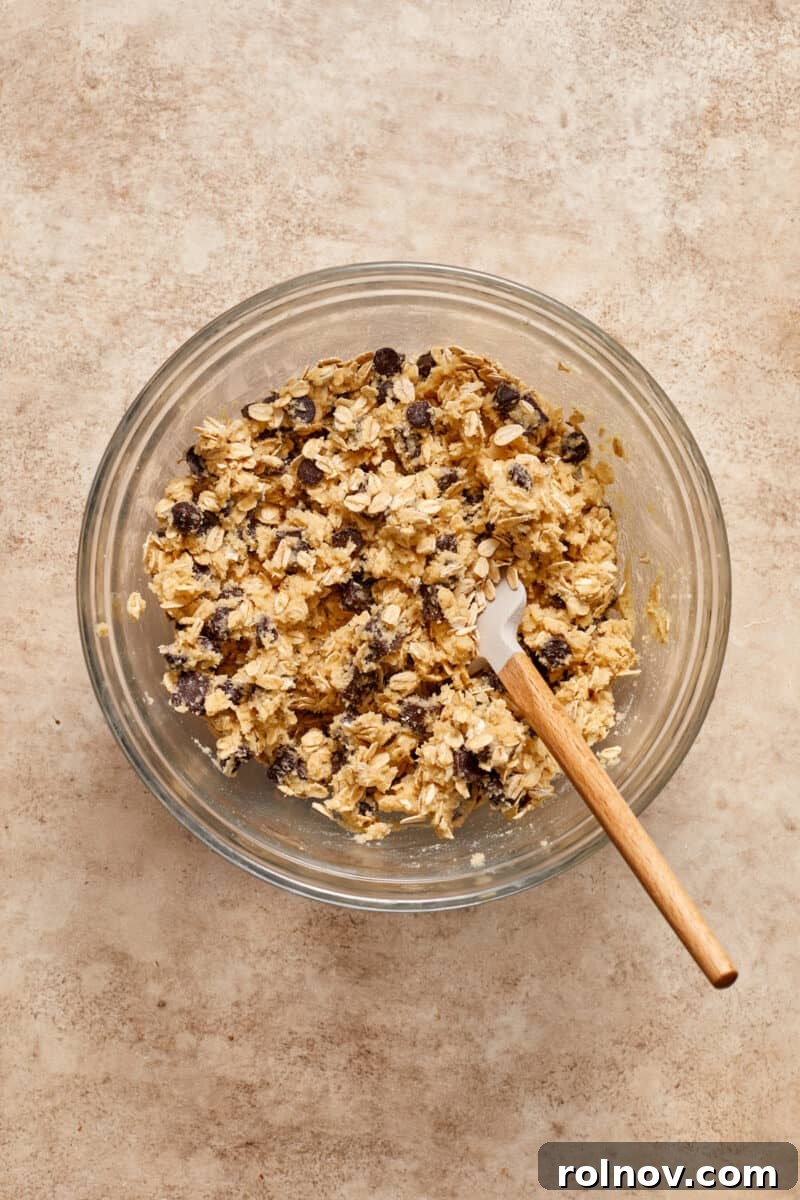 Almond & Oat Harmony Gluten-Free Chip Cookies 8 Smooth almond flour oatmeal chocolate chip cookie dough resting in a clear glass mixing bowl, with a spatula placed inside.