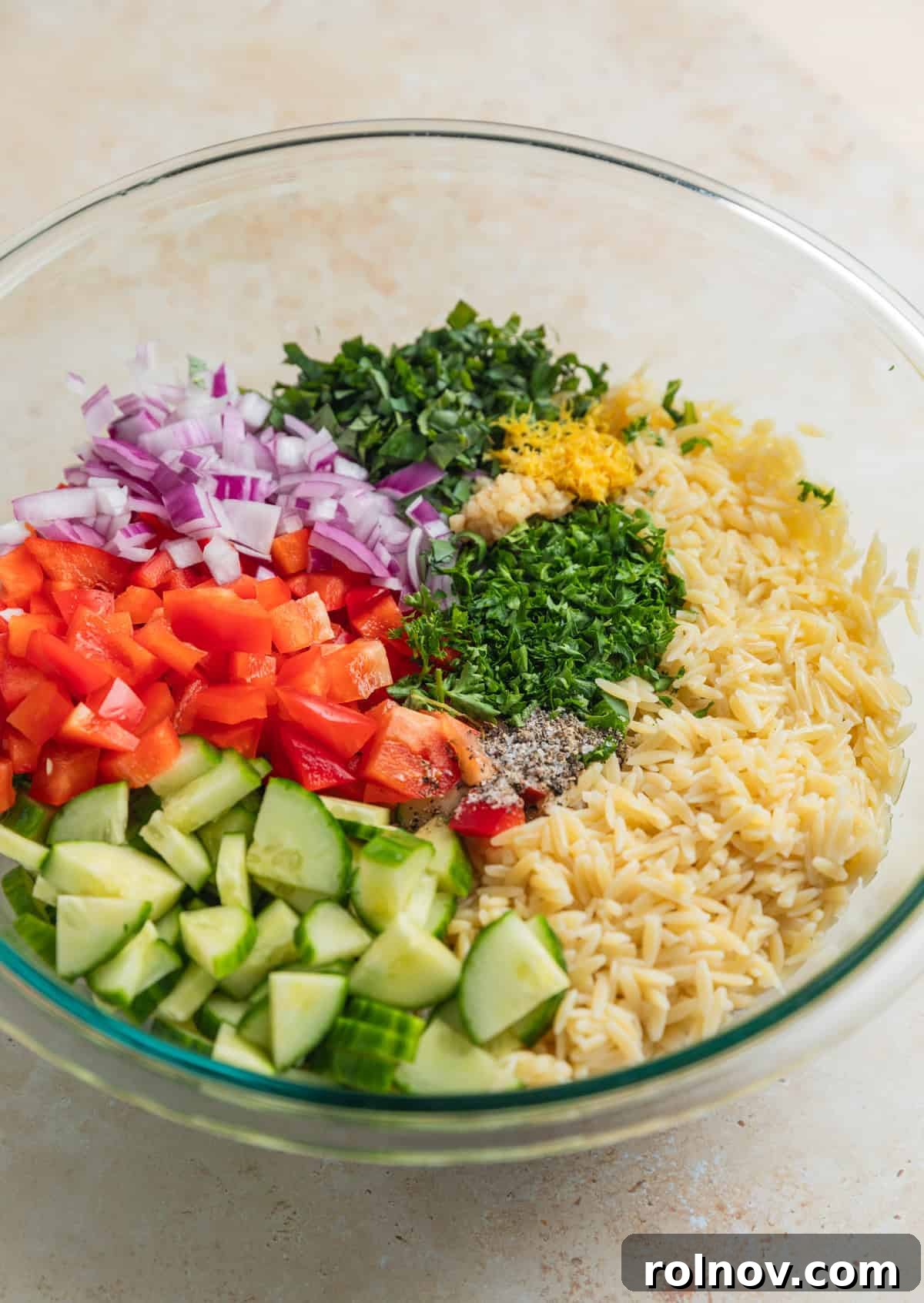 Bright & Effortless Lemon Orzo Salad with Feta 5 Orzo, lemon zest, cucumber, peppers and other ingredients in glass mixing bowl before mixing.