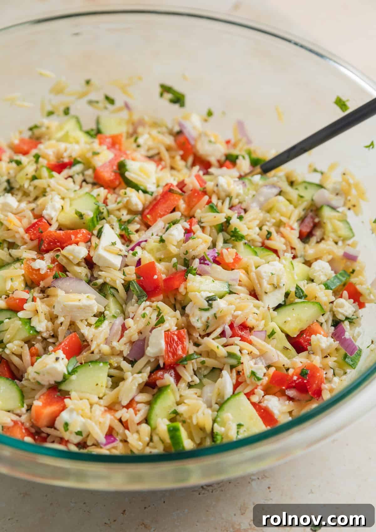 Bright & Effortless Lemon Orzo Salad with Feta 8 Prepared lemon orzo salad with spoon in glass mixing bowl, ready for serving.