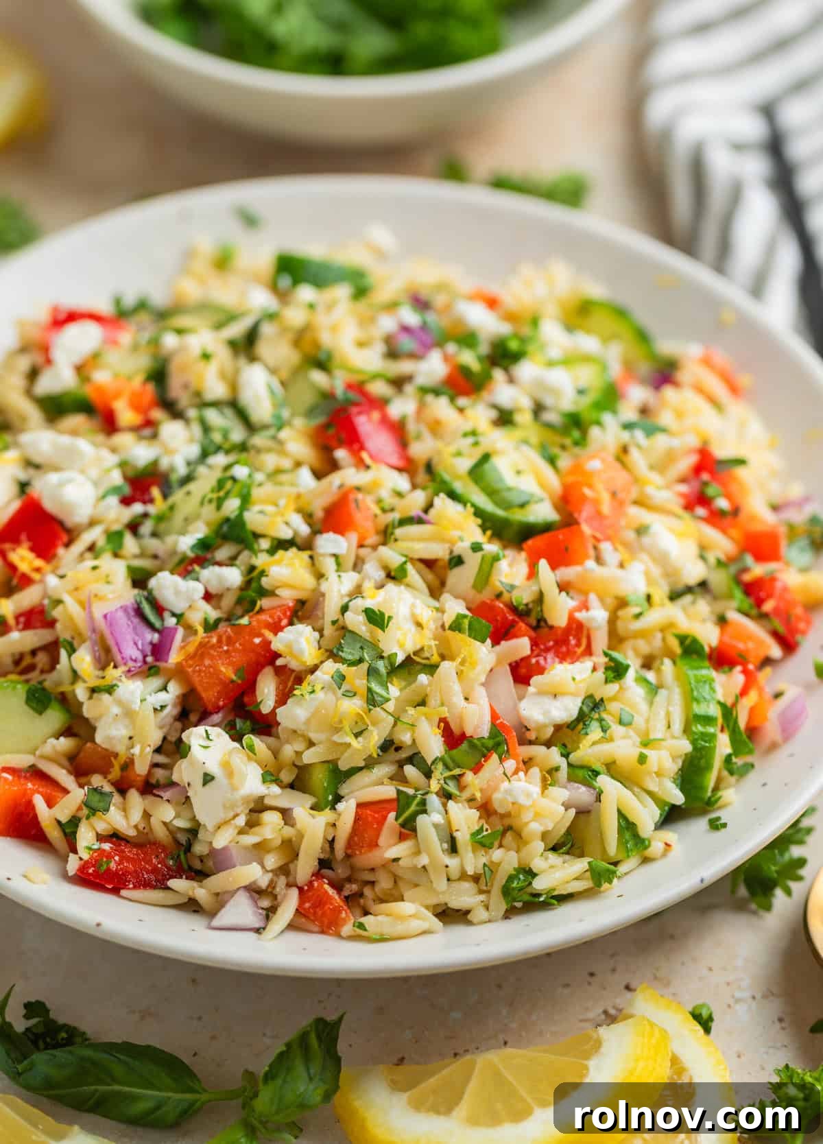 Bright & Effortless Lemon Orzo Salad with Feta 9 Orzo salad with feta, basil, parsley, cucumbers and more in white bowl, ready to be served.