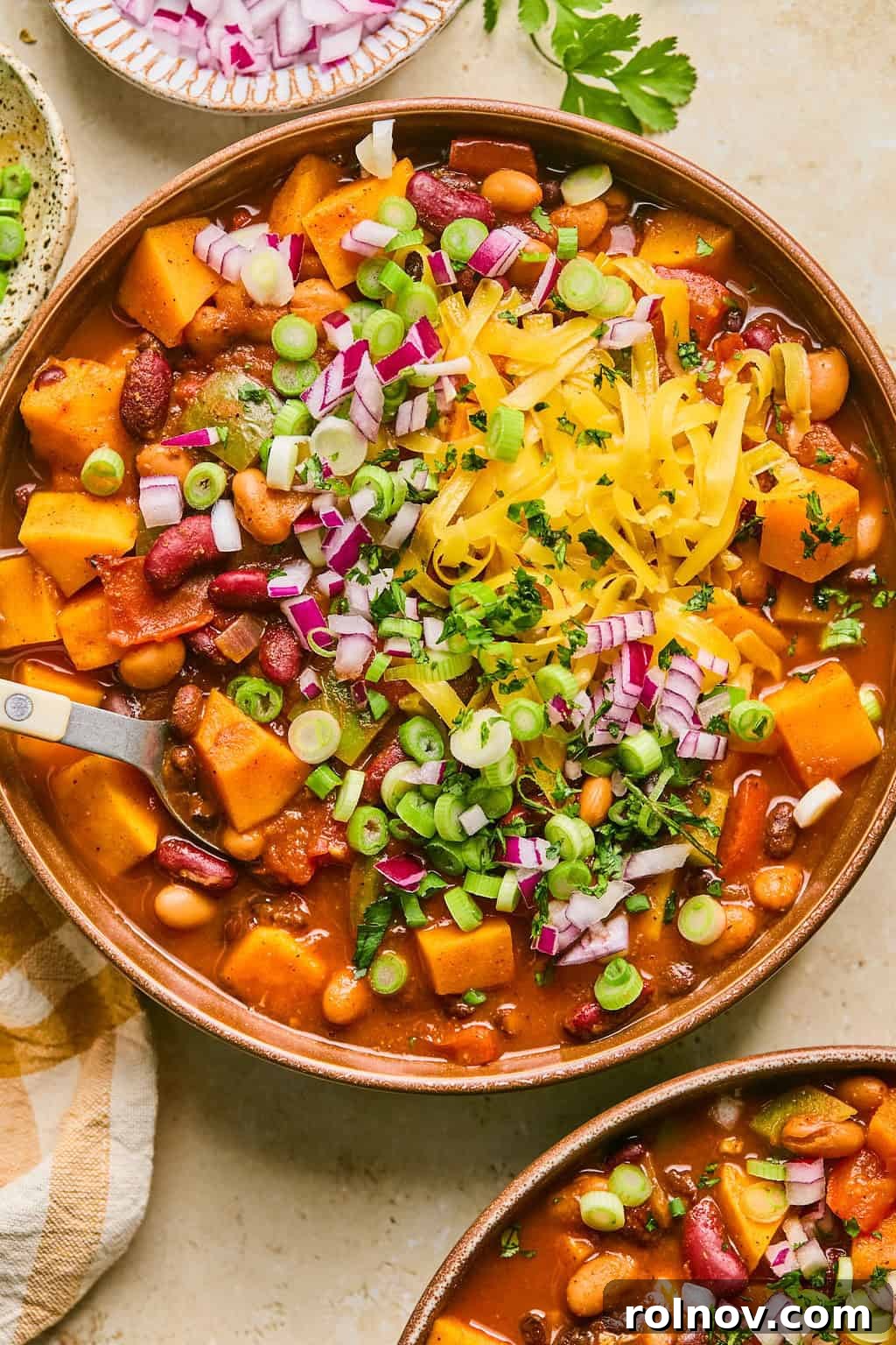 Bowl of vibrant butternut squash chili, generously garnished with shredded cheese, freshly chopped red onion, and bright green cilantro.