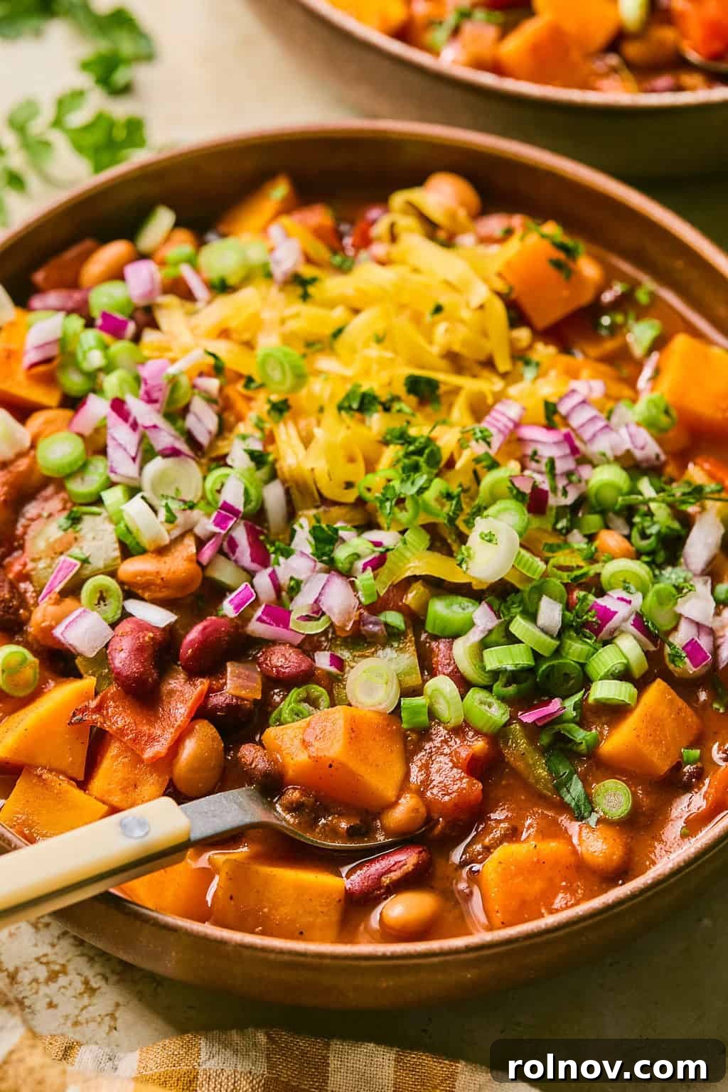 A close-up shot of a bowl of butternut squash chili, garnished with a sprinkle of shredded cheese and finely chopped onions, with a spoon ready for enjoyment.