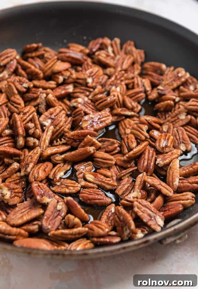 Pecans being stirred in a skillet with a clear maple syrup mixture, showing the initial coating process on the stovetop.