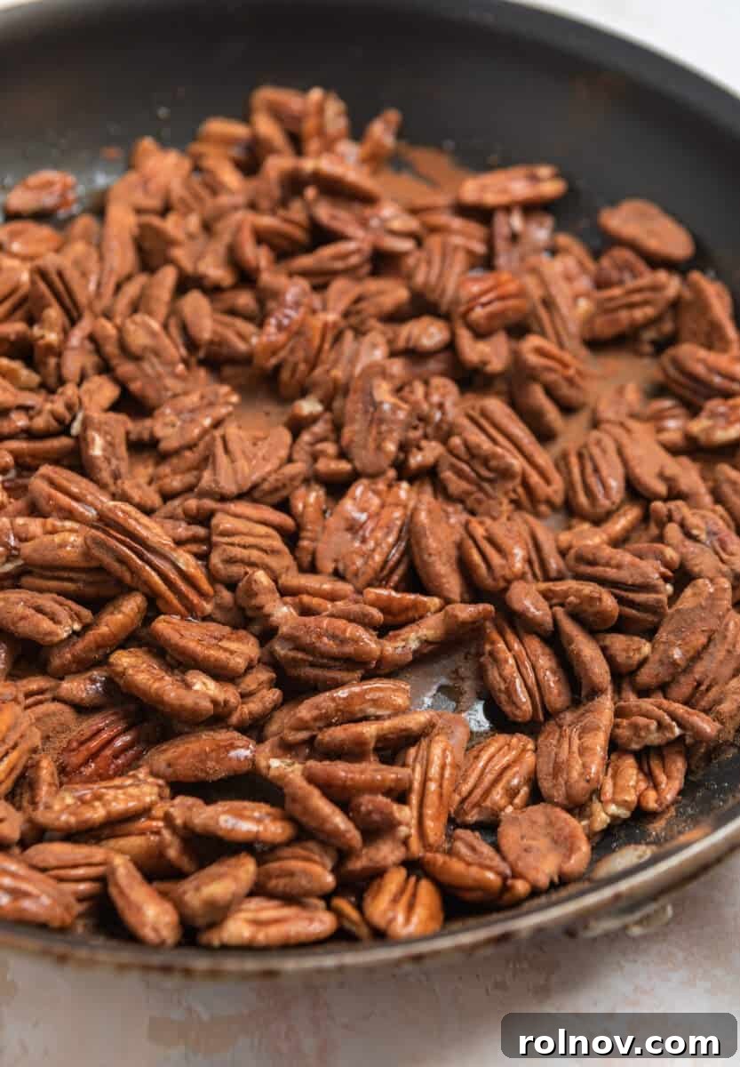 Ground cinnamon being sprinkled evenly over the maple-coated pecans in a skillet, creating a fragrant layer before further stirring.