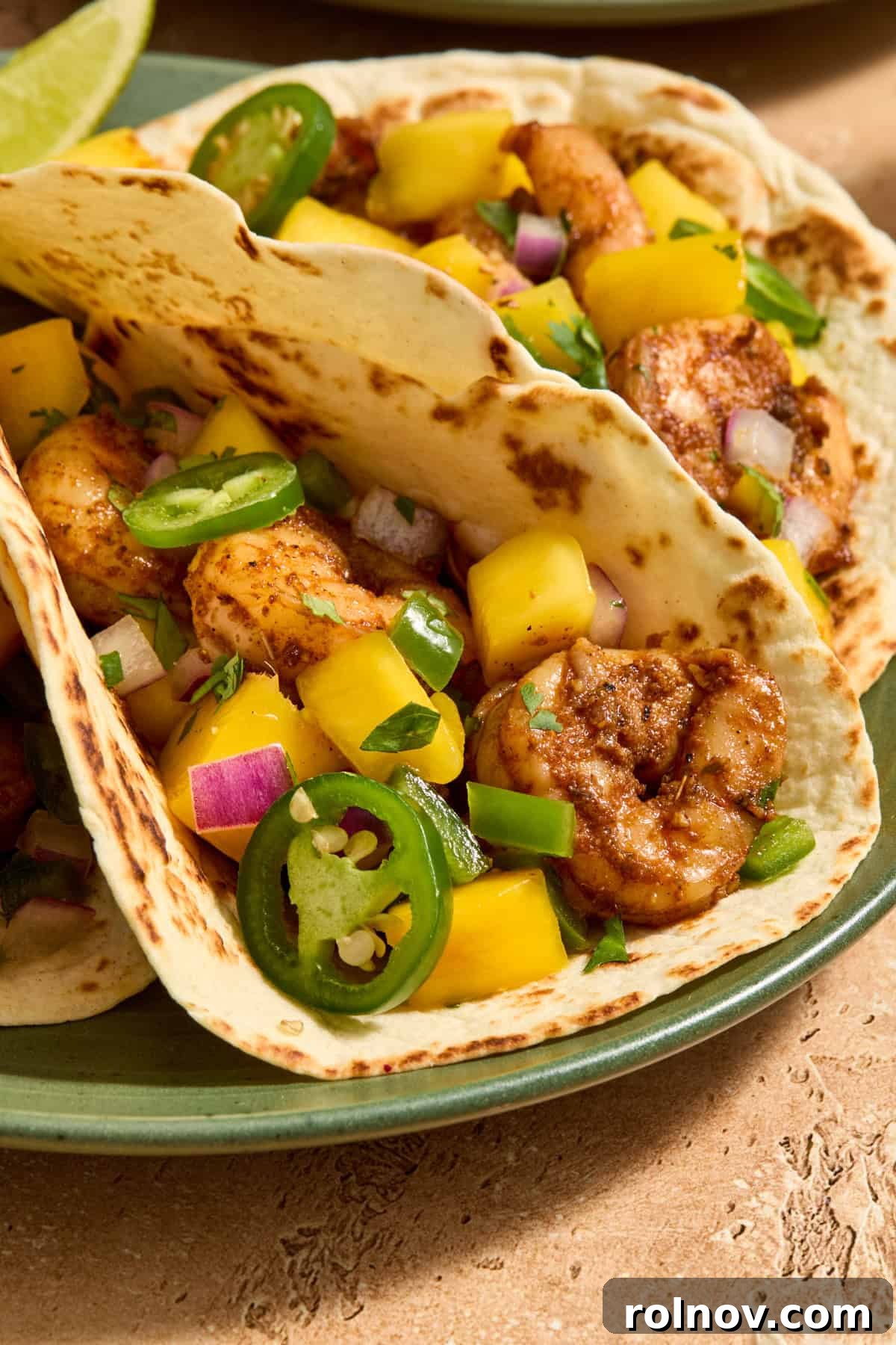 Tropical Shrimp Tacos with Mango Salsa 3 Close-up view of two spicy shrimp tacos adorned with fresh mango salsa, vibrant cilantro, and thin slices of jalapeños on a modern plate.