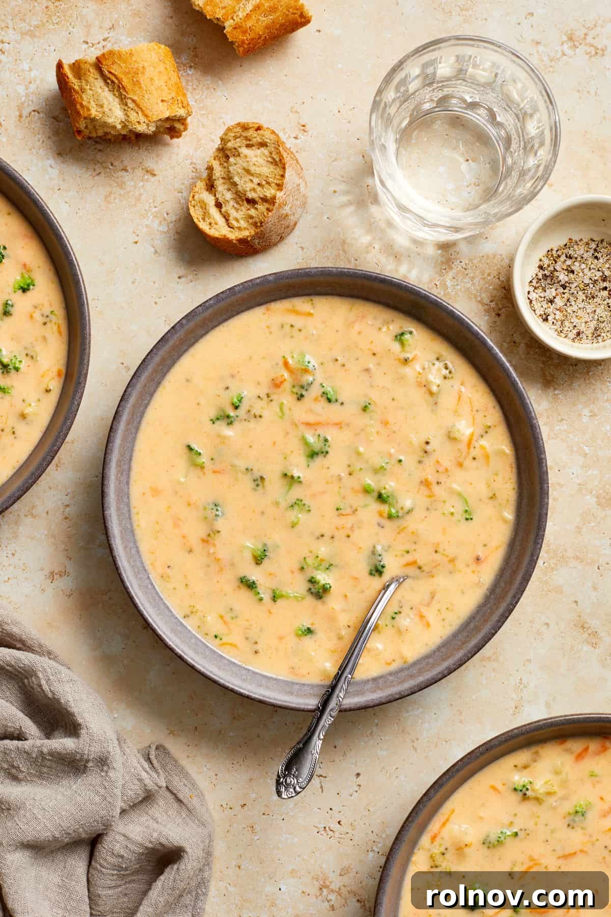 Creamy Instant Pot Broccoli Cheddar Soup 11 A beautifully set table features a bowl of steaming broccoli and cheese soup, accompanied by a glass of water, an assortment of crusty bread pieces, and smaller bowls, creating a warm and inviting meal scene.