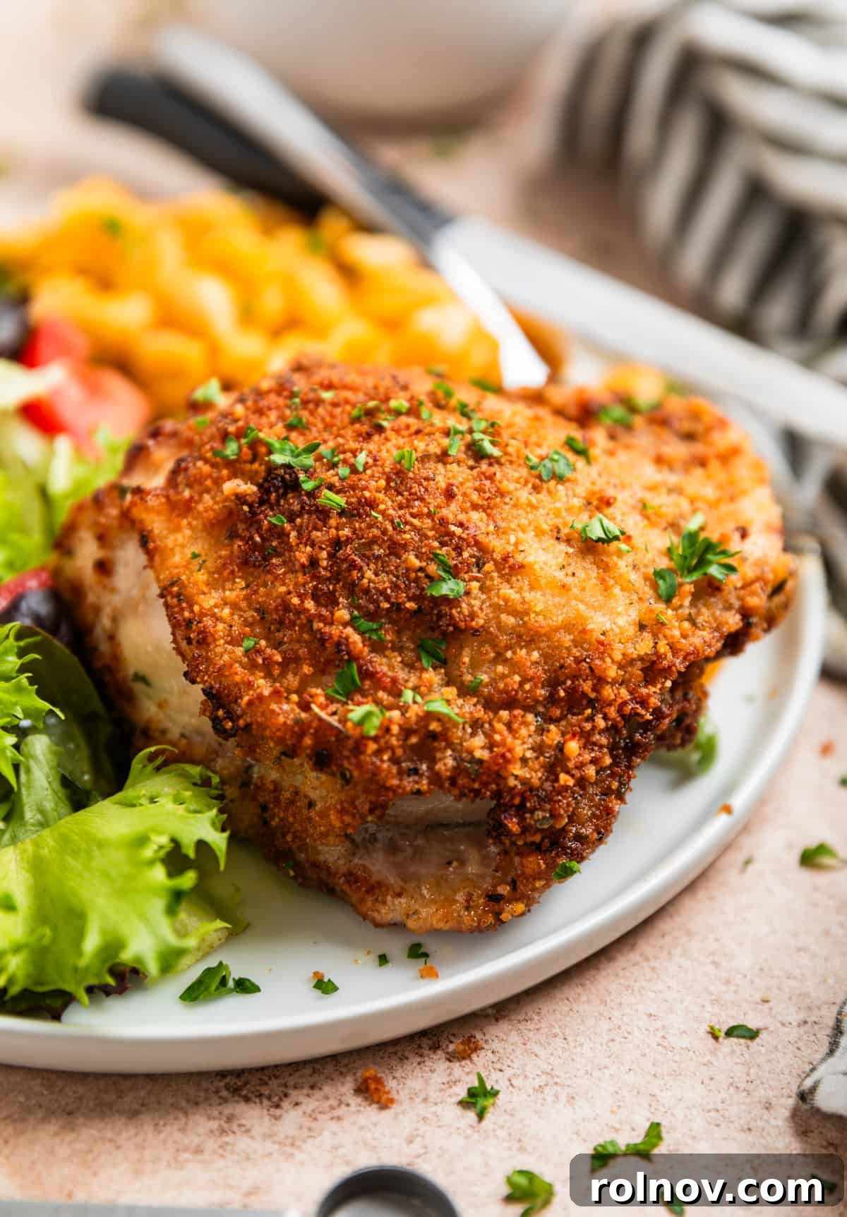 Crispy Parmesan Air Fryer Chicken Thighs 2 Crispy breaded parmesan air fryer chicken thigh on a white plate, garnished with chopped parsley, served alongside creamy macaroni and cheese and a fresh green salad.