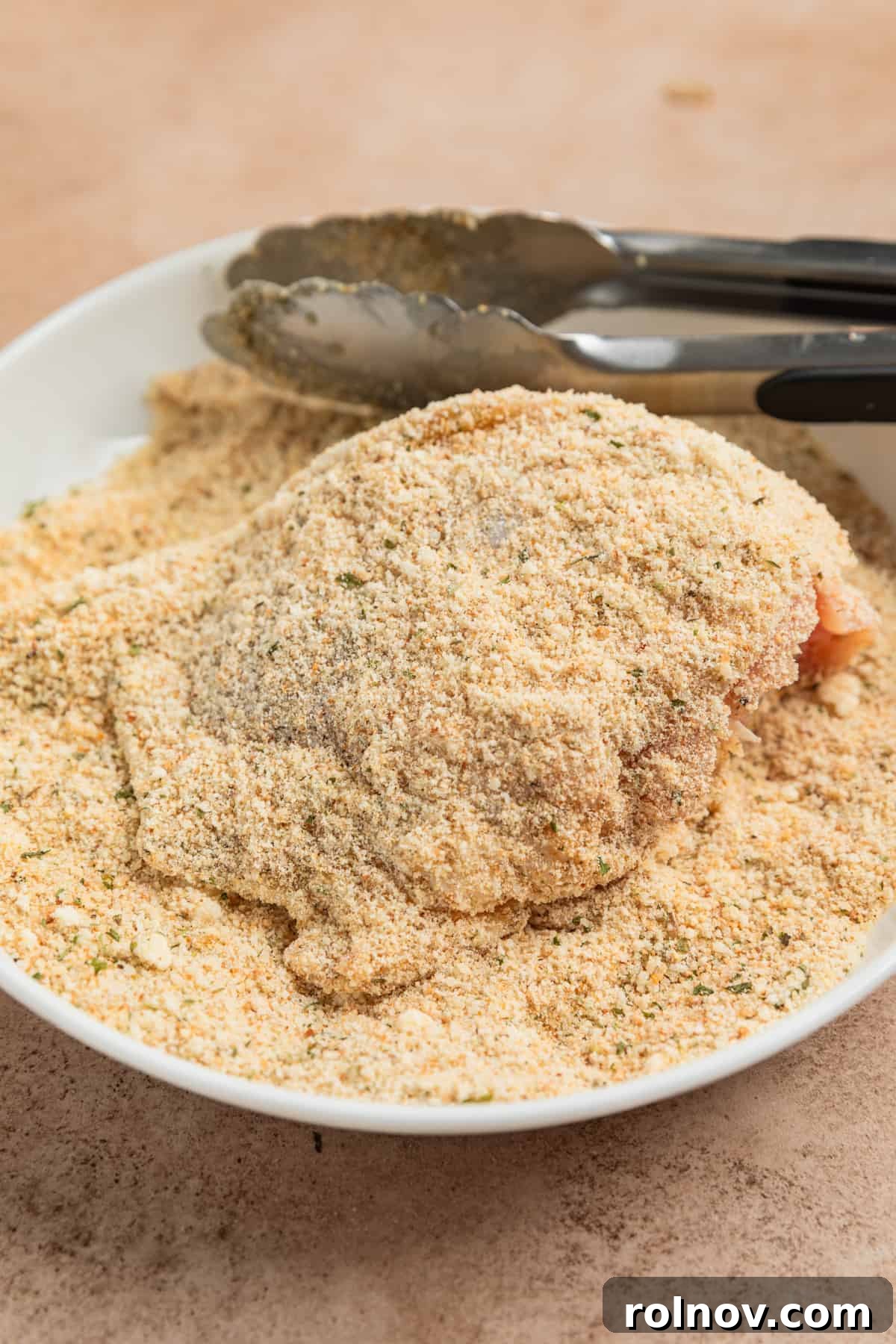 Crispy Parmesan Air Fryer Chicken Thighs 5 A chicken thigh being coated with tongs in a shallow dish filled with a mixture of breadcrumbs and grated Parmesan cheese, ensuring full coverage.