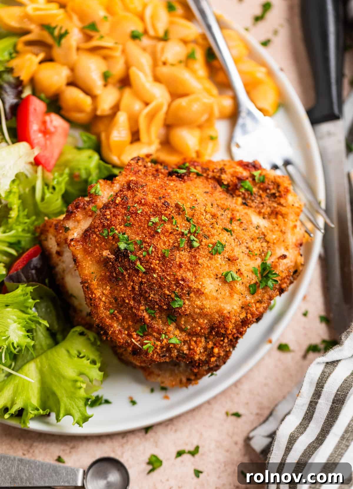 Crispy Parmesan Air Fryer Chicken Thighs 8 An overhead view of a golden-brown, crispy parmesan crusted chicken thigh on a white plate, served with a fork, alongside a creamy mac and cheese portion and a fresh garden salad.