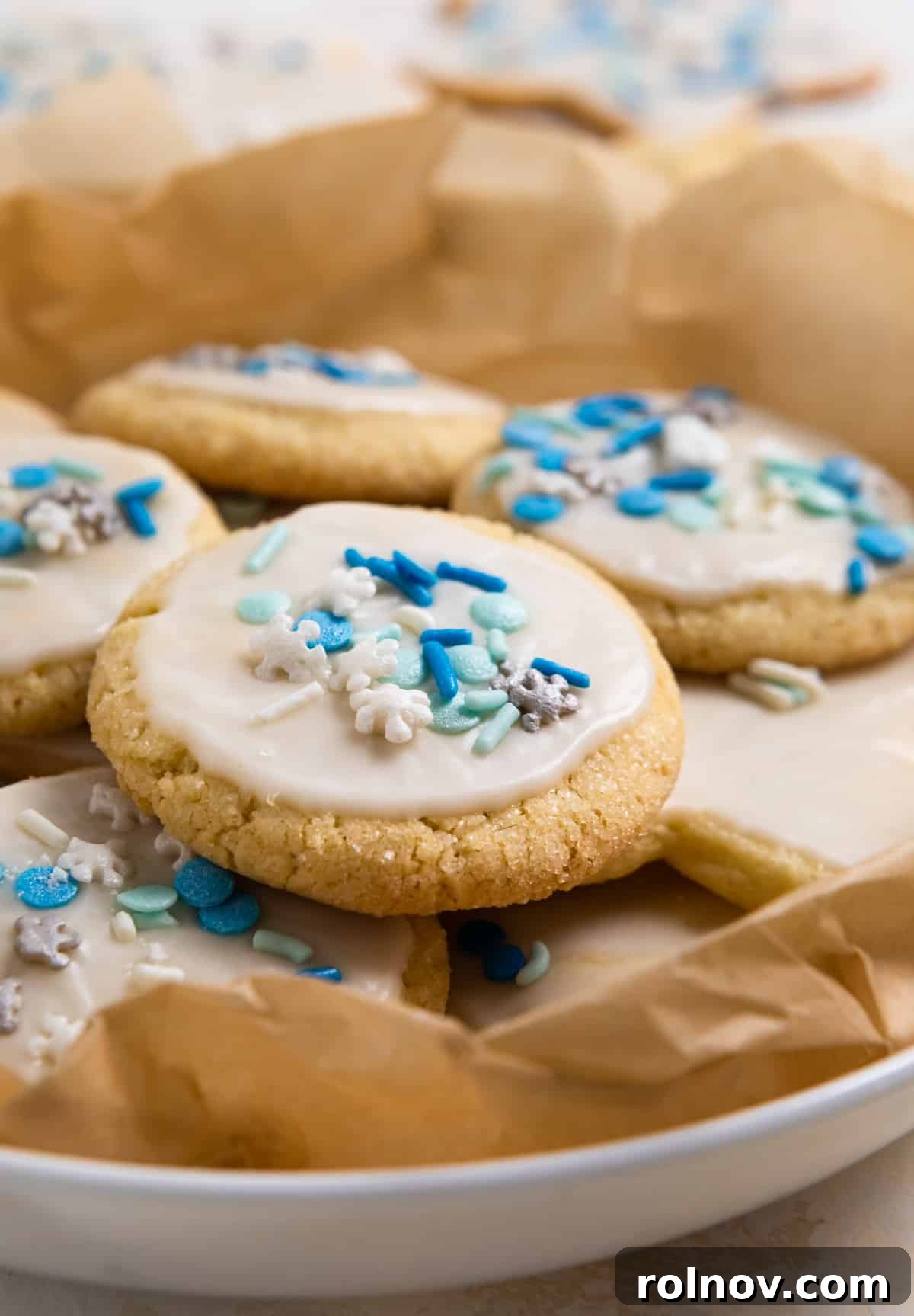 Plate of perfectly baked eggless sugar cookies, elegantly adorned with a pristine white glaze and festive blue sprinkles, resting on parchment paper.