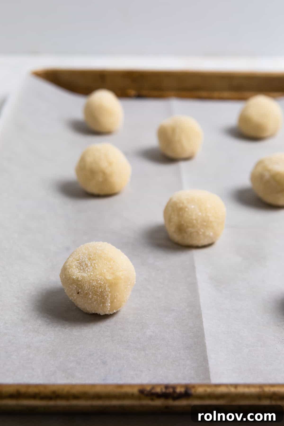 A baking sheet lined with parchment paper, displaying neatly rolled sugar cookie dough balls, coated in sugar and spaced perfectly for baking.