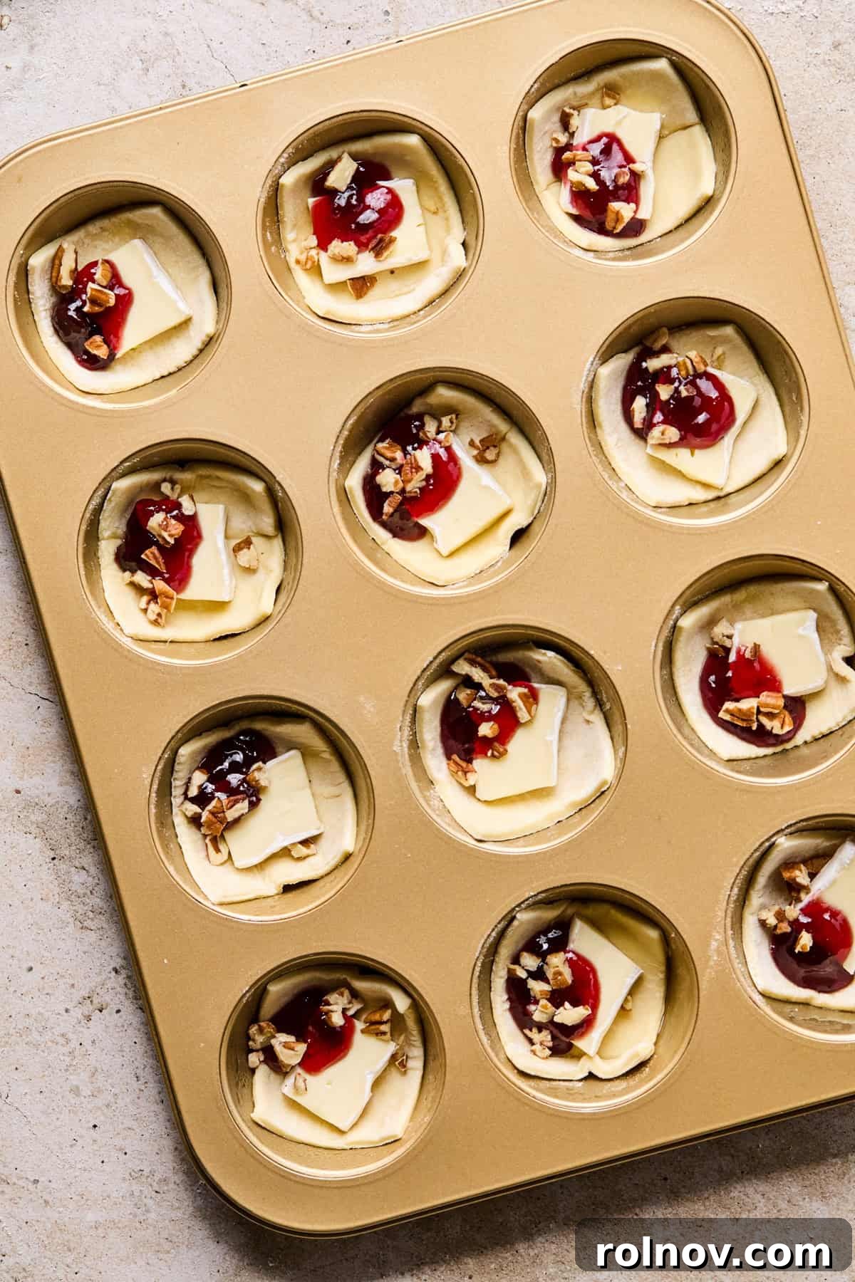 Golden Brie Pastry Parcels 6 Puff pastry brie bites in a muffin tin, topped with jam and pecans, ready for baking.
