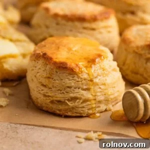Simple honey biscuits on tan parchment with honey drizzling over top with a honey dipper stick beside it.