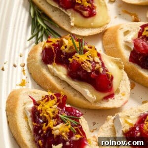Festive cranberry-topped brie crostini artfully arranged on a plate, garnished with fresh rosemary and bright orange zest.