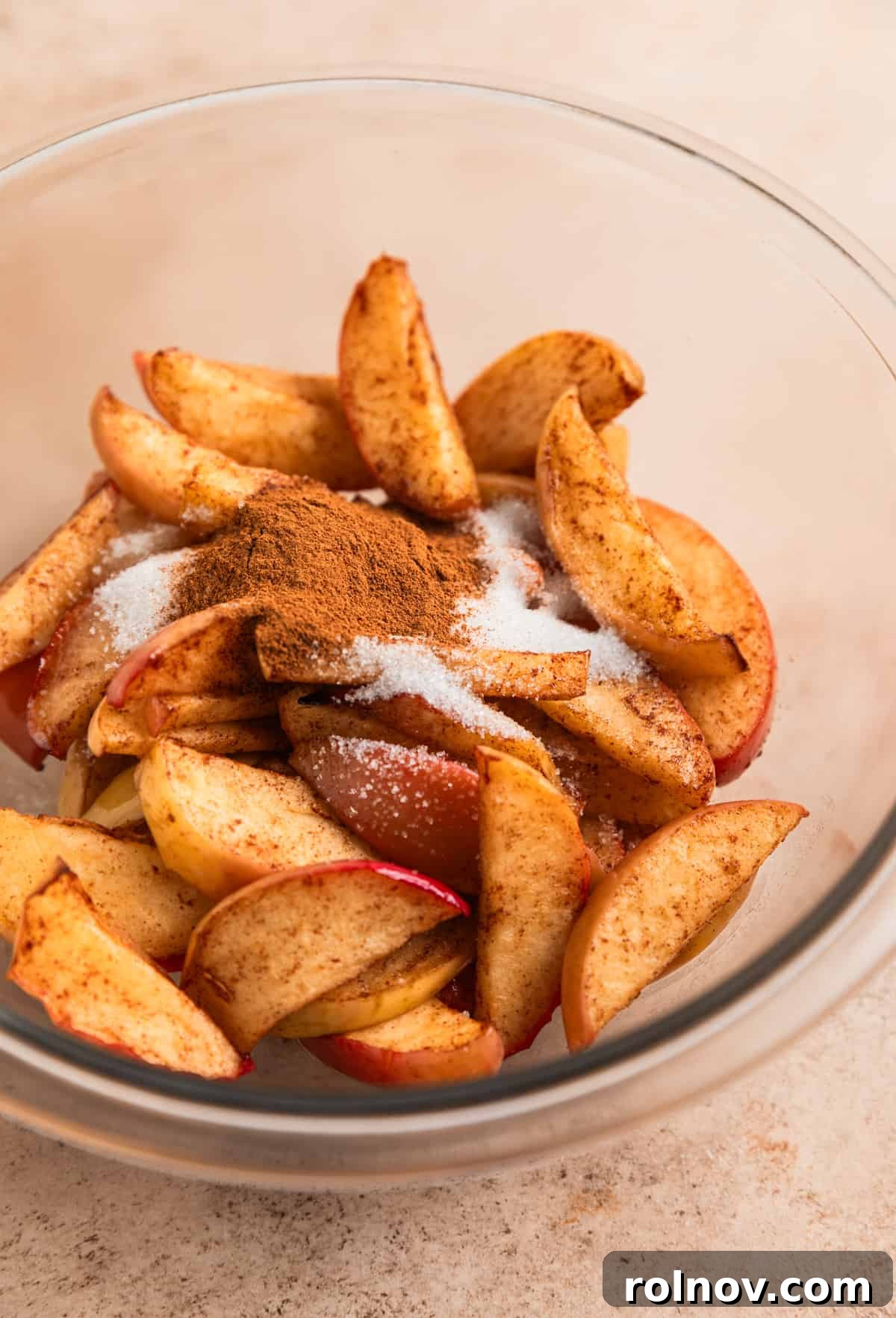 Air Fryer Cinnamon Apple Slices 6 Air fryer cinnamon apples in a glass mixing bowl, with melted butter, sugar, and cinnamon being poured on top.