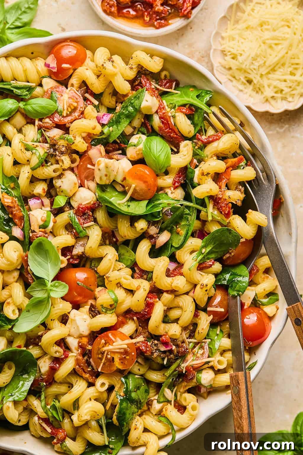 A vibrant bowl of Sun-Dried Tomato Pasta Salad, garnished with fresh basil, featuring rigatoni pasta, spinach, cherry tomatoes, and mozzarella pearls. A spoon and fork are propped in the bowl, inviting a taste.
