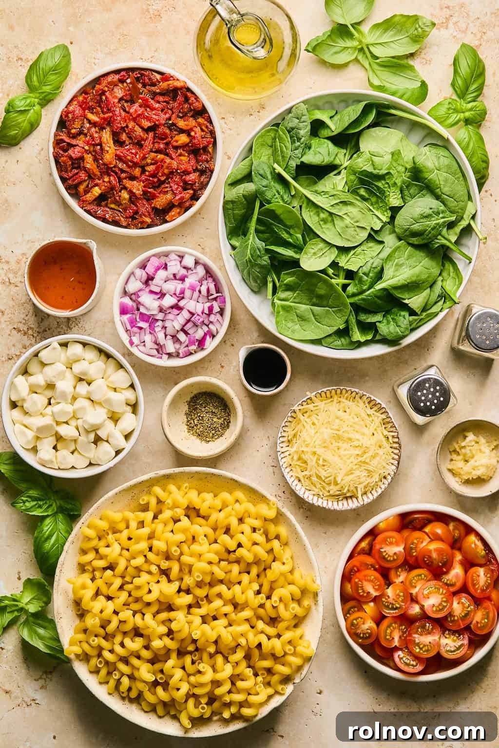 An array of fresh ingredients for sun-dried tomato pasta salad laid out on a surface, including fresh spinach, uncooked pasta, a wedge of parmesan cheese, a bottle of olive oil, and jars of sun-dried tomatoes.