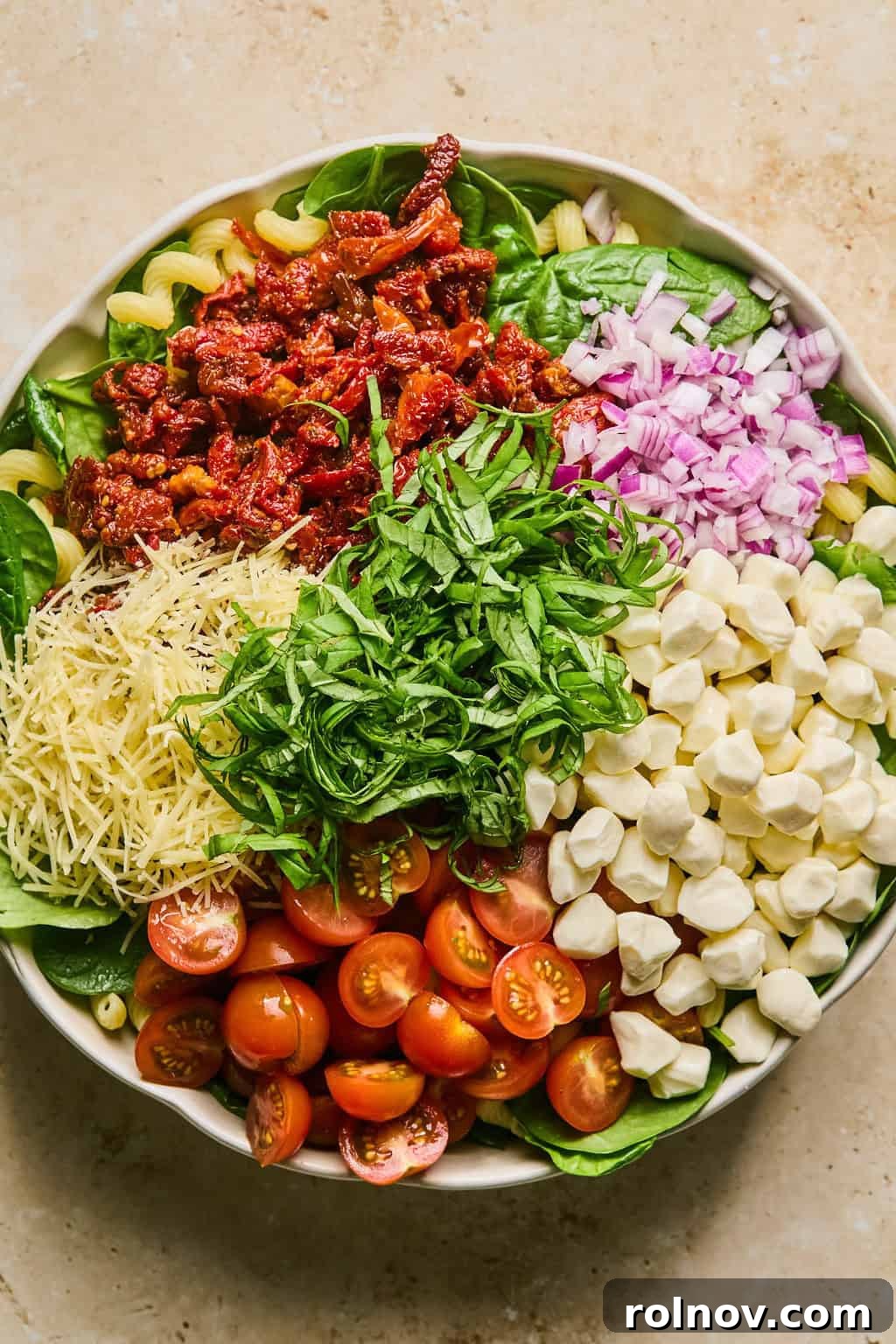 A large bowl of pasta and spinach topped with halved cherry tomatoes, finely diced red onion, mozzarella pearls, and shredded Parmesan cheese.
