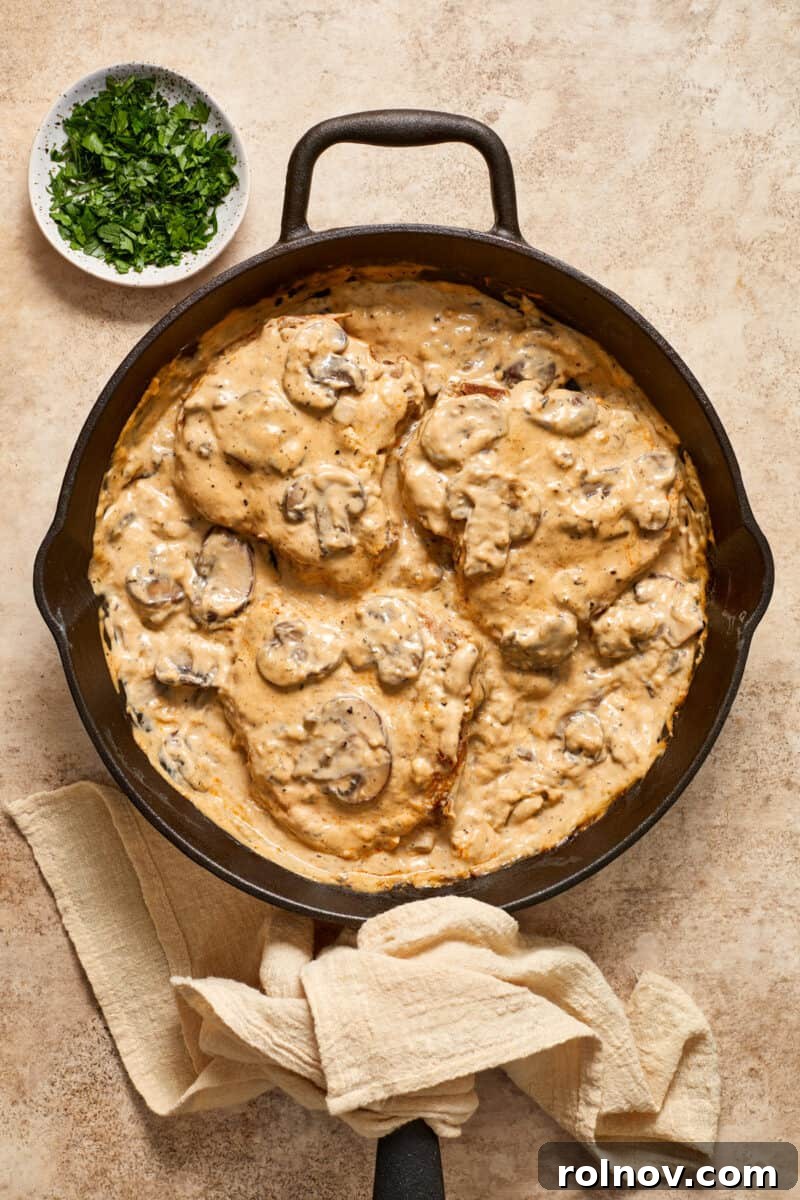 Velvety Mushroom Pork Chops 12 Cast iron skillet with creamy mushroom bone in pork chops and bowl of parsley beside it.