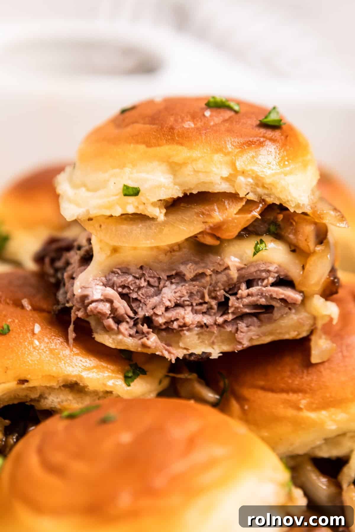 Ultimate French Dip Sliders 2 French dip slider with roast beef, provolone, onion and mushroom stacked on other sandwiches, ready to be served.