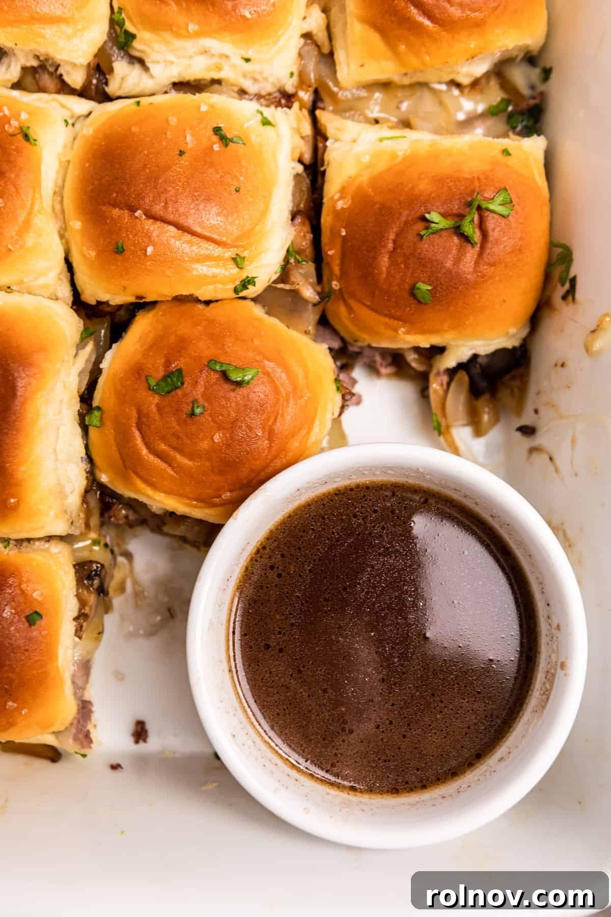 Ultimate French Dip Sliders 11 Overhead view of au jus in a small white bowl, surrounded by perfectly baked French dip sliders.