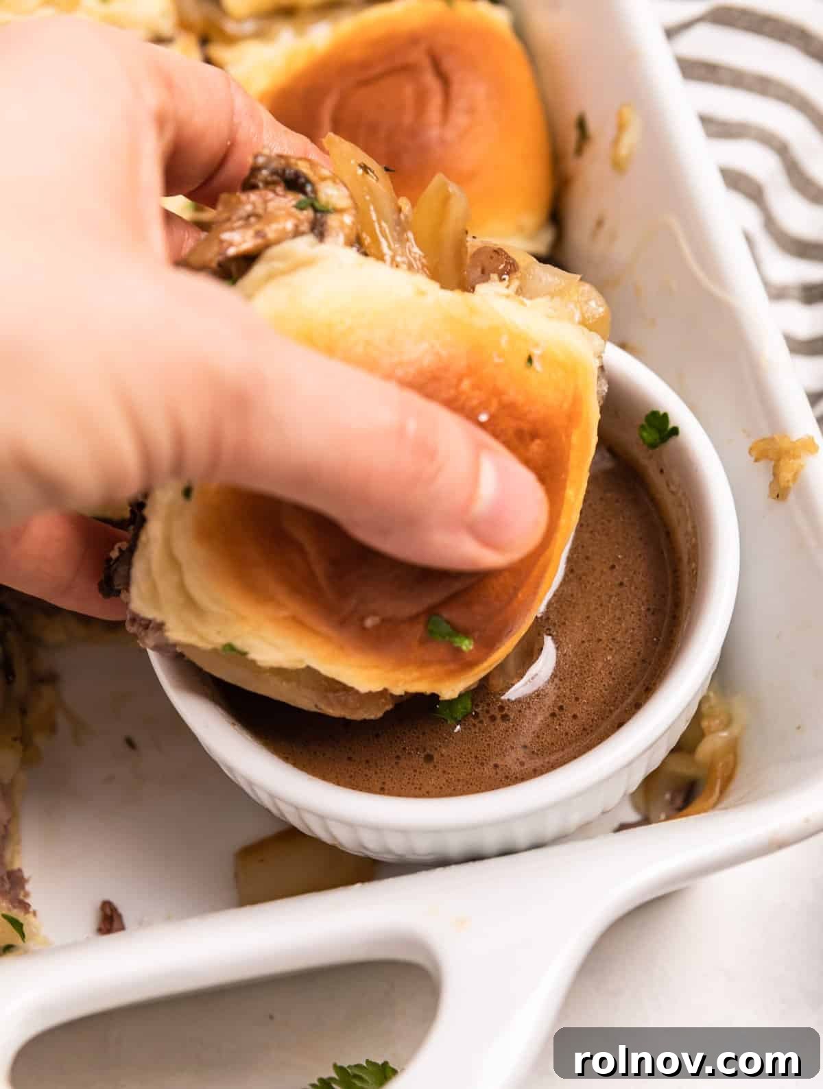 Ultimate French Dip Sliders 12 A single French dip slider, perfectly dipped into a small bowl of au jus, showing the delicious, moist interior.