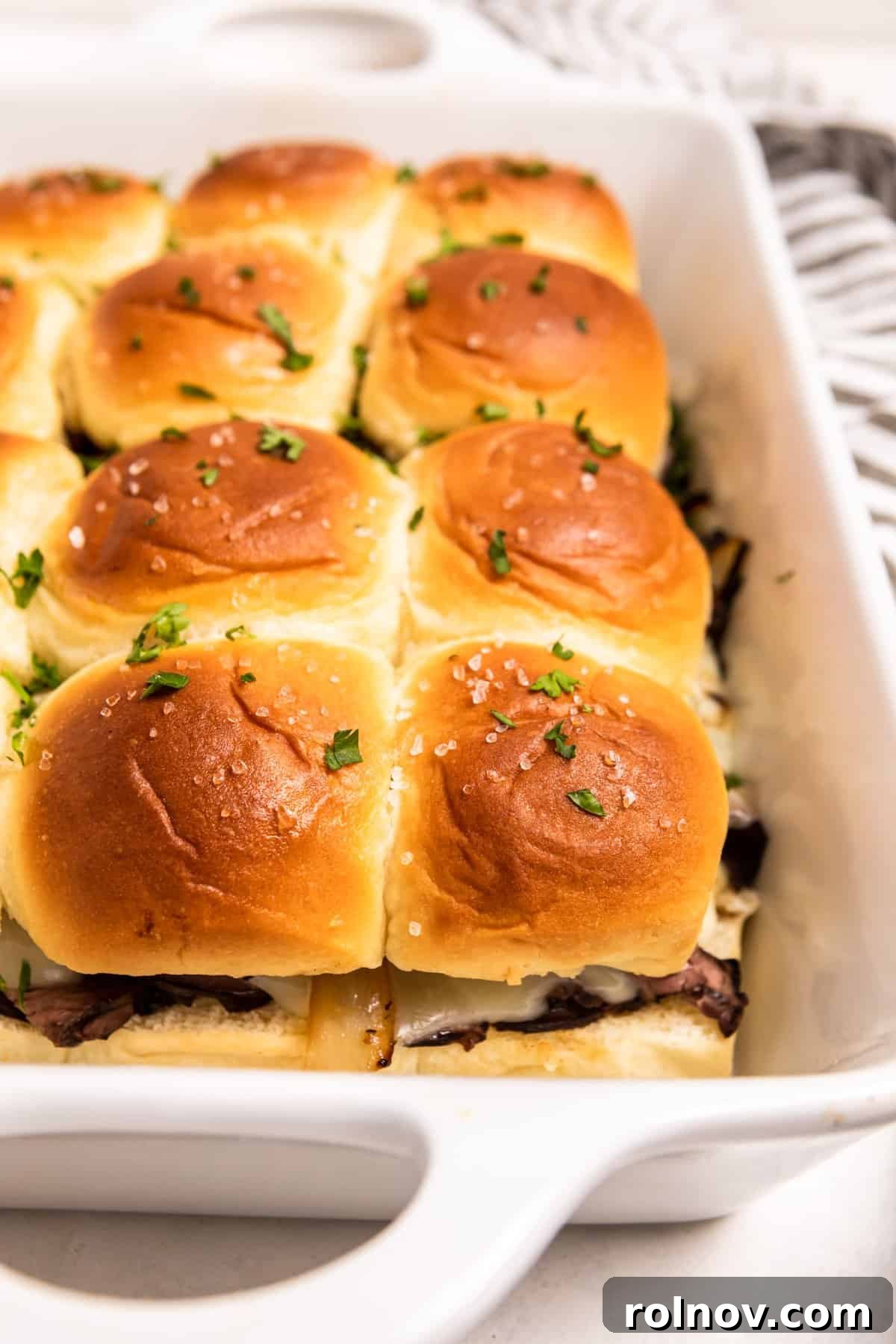 Ultimate French Dip Sliders 13 White baking dish filled with fresh French dip sliders, garnished with chopped parsley, ready for serving.