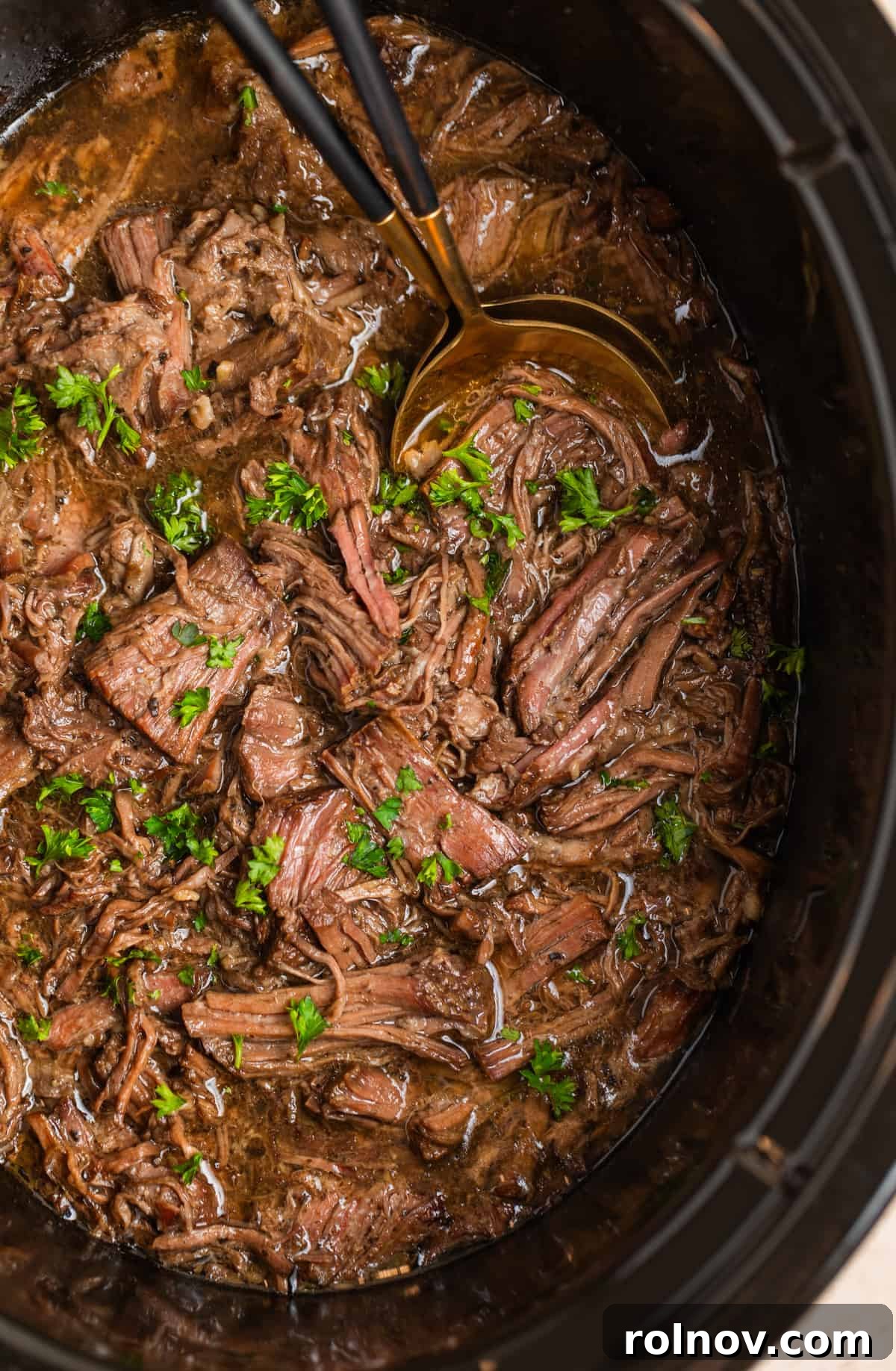 Slow cooker shredded beef in crockpot with serving utensils and chopped parsely.