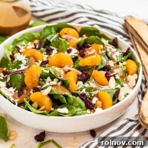 White salad bowl with spinach, cranberries, almonds, goat cheese, oranges and balsamic dressing drizzled on top.