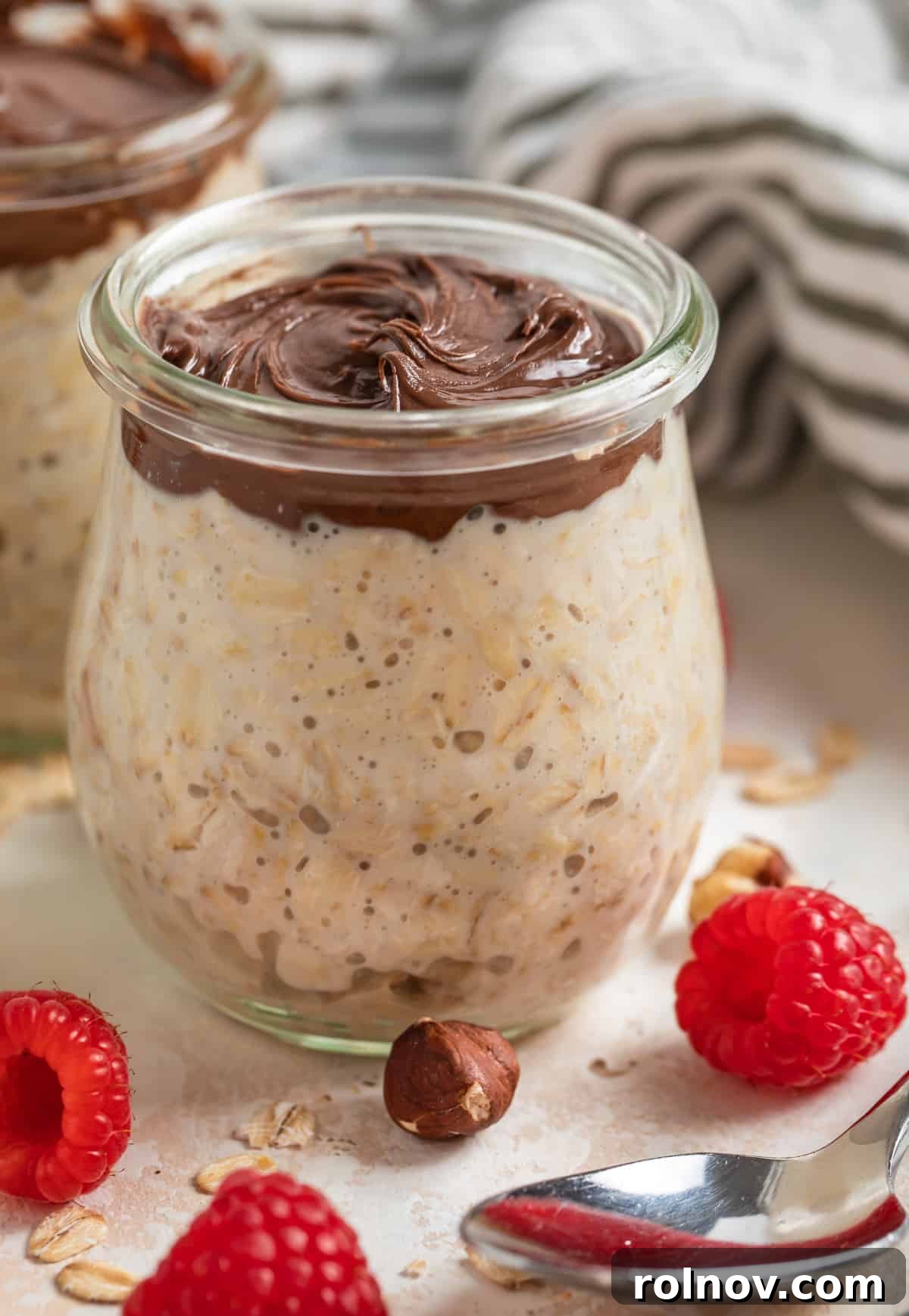 Nutella overnight oats in a stylish Weck jar, surrounded by scattered fresh raspberries and whole hazelnuts, highlighting the rich and inviting texture.