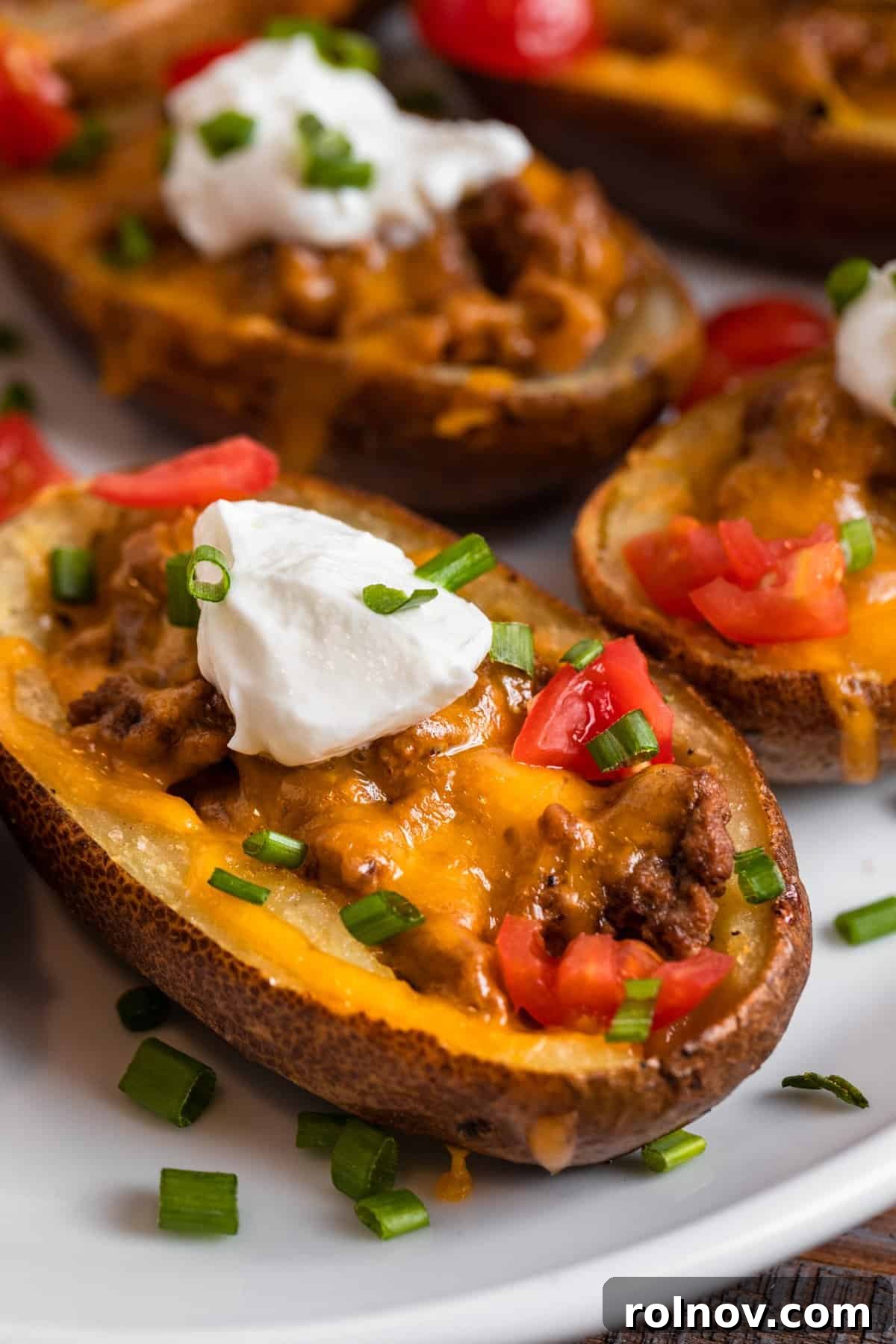Cheesy Burger Potato Boats 2 Cheeseburger stuffed potato skins on white plate topped with sour cream, chives and tomatoes.