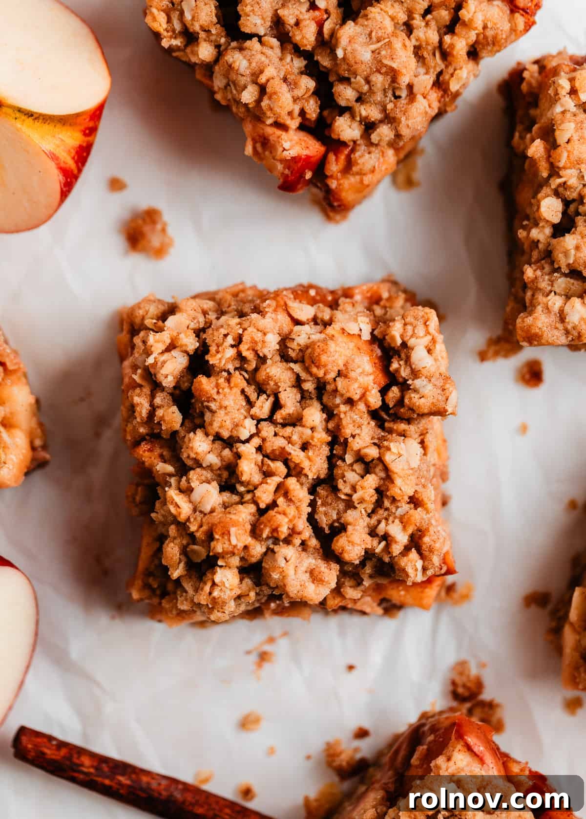 Golden Apple Crumble Bars 7 Overhead view of a single apple crumb bar with a cinnamon stick and fresh apple slices arranged beside it on parchment paper.
