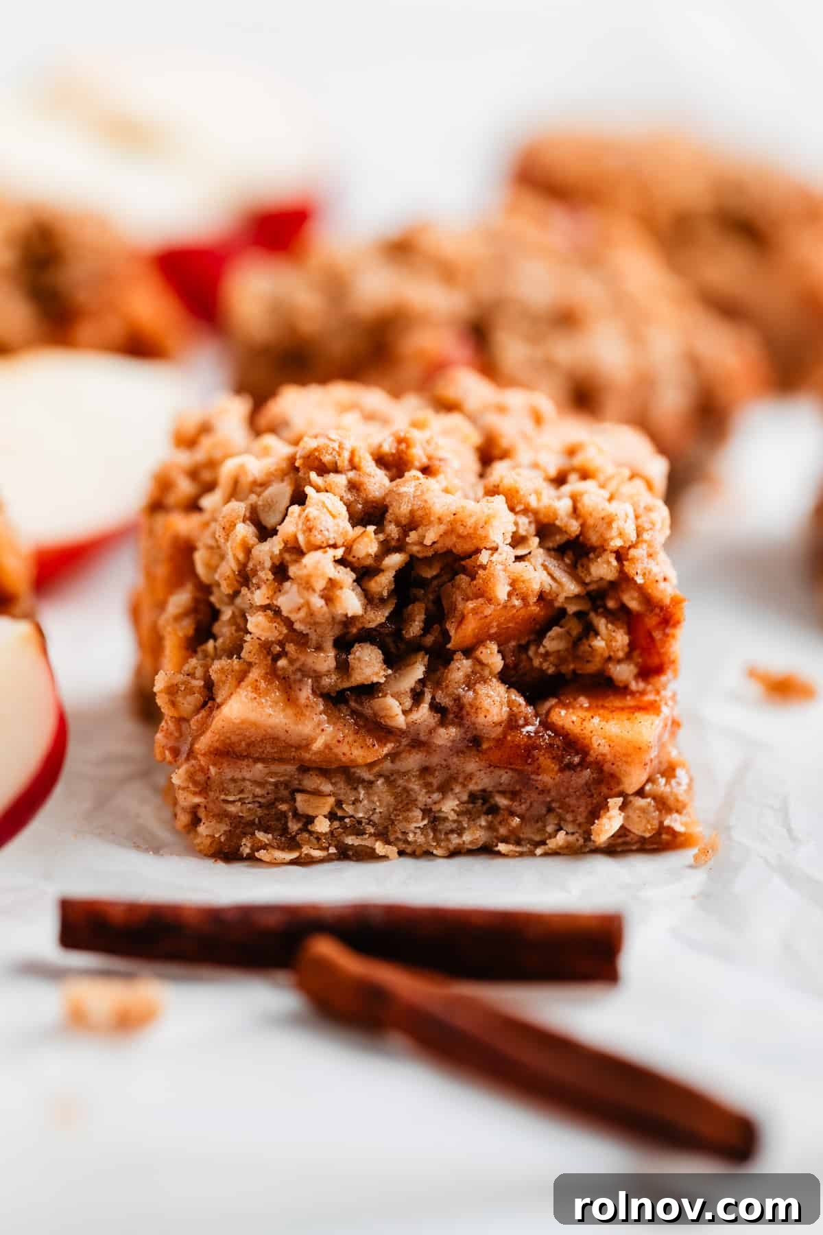 Golden Apple Crumble Bars 8 A slice of layered apple crumble bar resting on parchment paper, surrounded by cinnamon sticks for garnish.