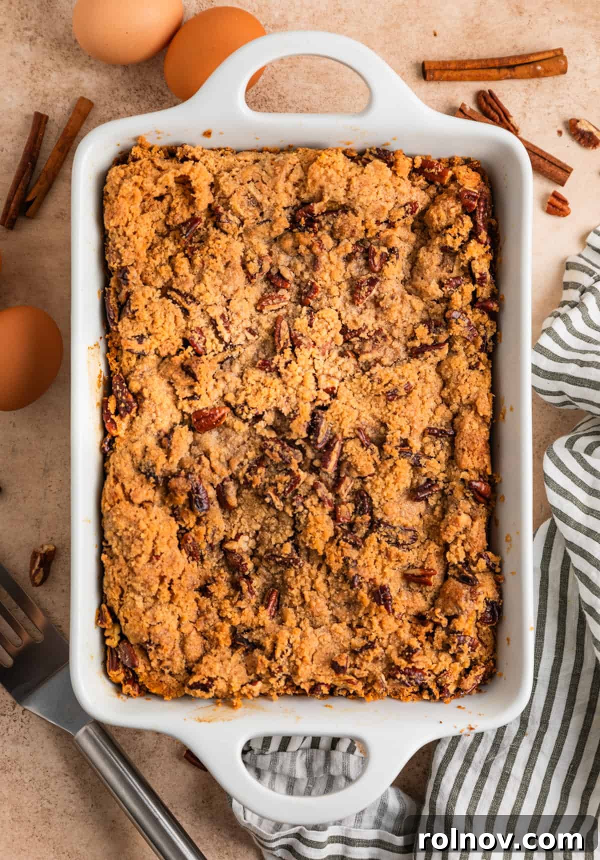 Pumpkin Spice French Toast Bake 10 White pan with streusel topped baked French toast with striped linen, eggs and spatula beside the dish.