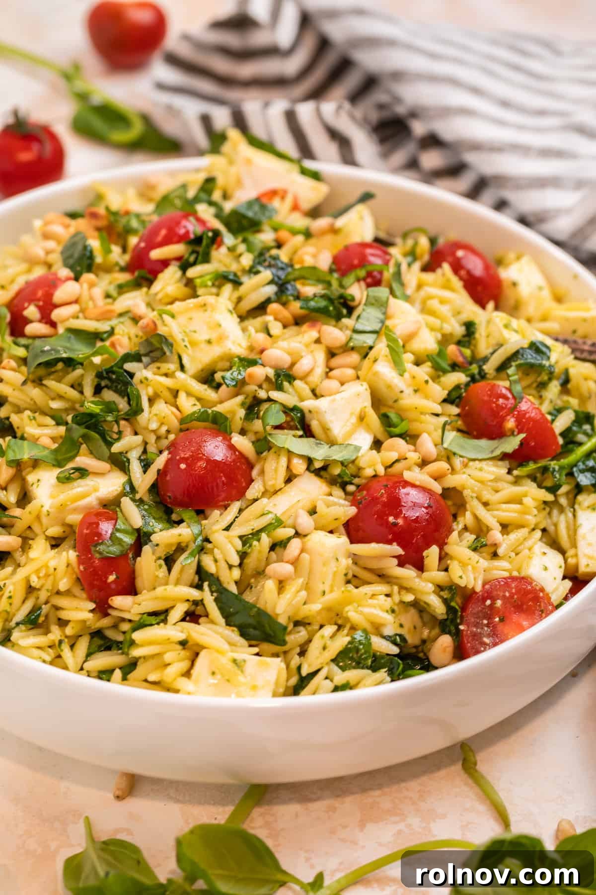 Emerald Orzo Pesto Salad 3 Orzo salad with pesto, mozzarella, tomatoes and chopped spinach in white bowl with towel beside it.