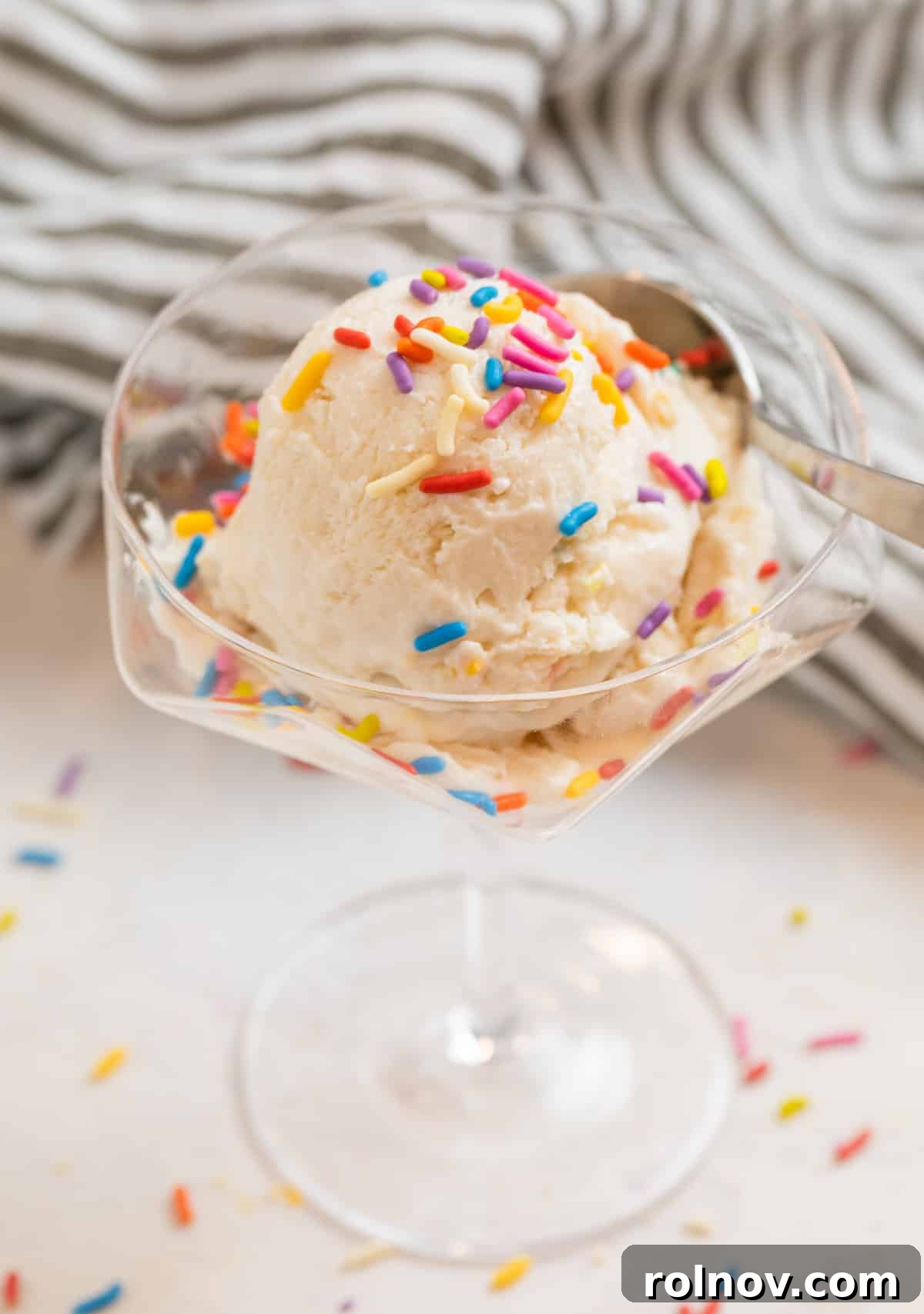 Cottage Cream Dream 12 A single scoop of classic vanilla cottage cheese ice cream in a clear glass dessert dish, garnished with rainbow sprinkles, with a spoon nearby.