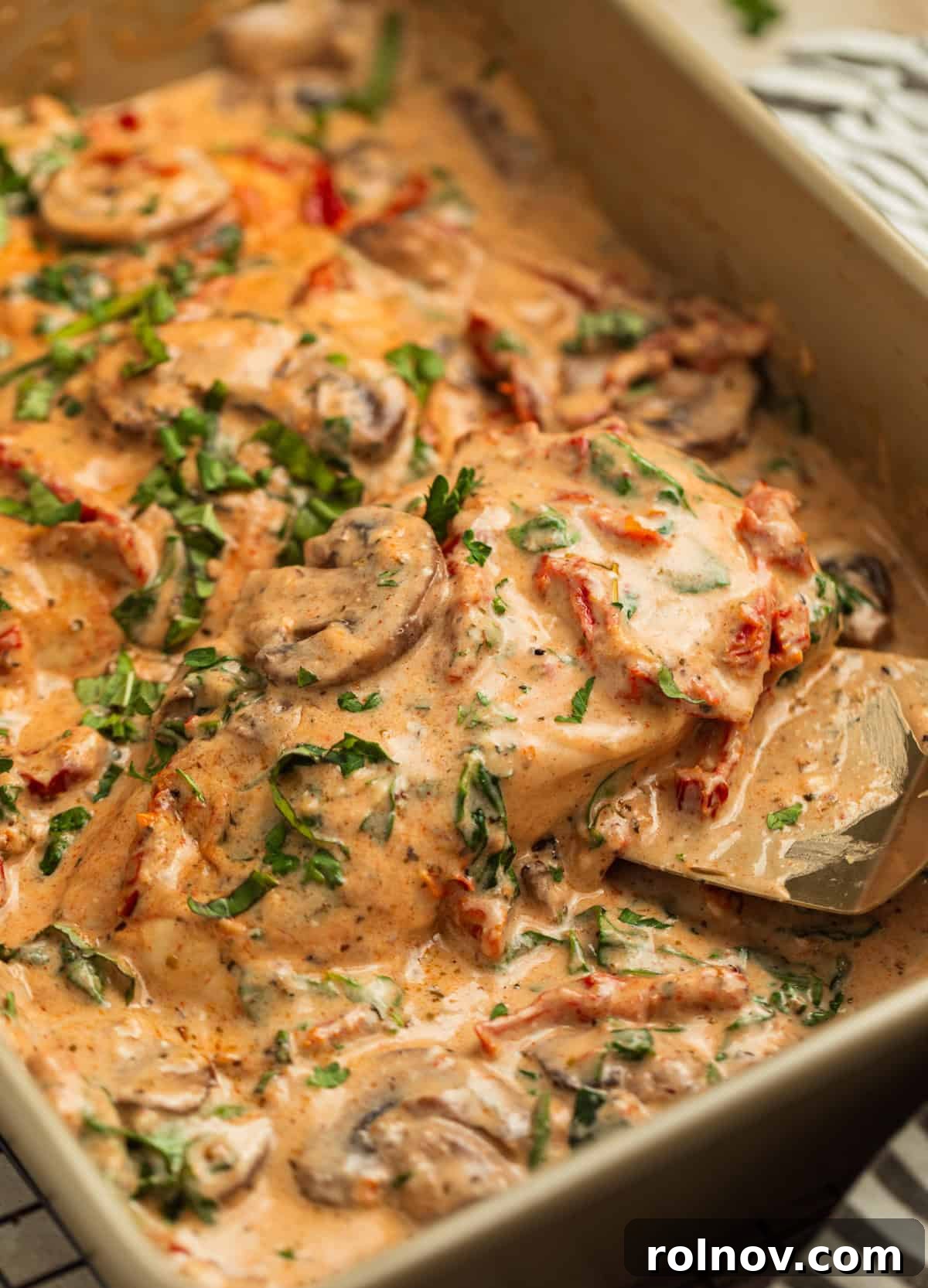 Savory Tuscan Chicken Bake 2 Pan with golden-baked Tuscan chicken breast, rich creamy sauce, mushrooms, spinach, and sun-dried tomatoes. A spatula is lifting a piece of chicken, showcasing its juiciness and the delicious sauce.