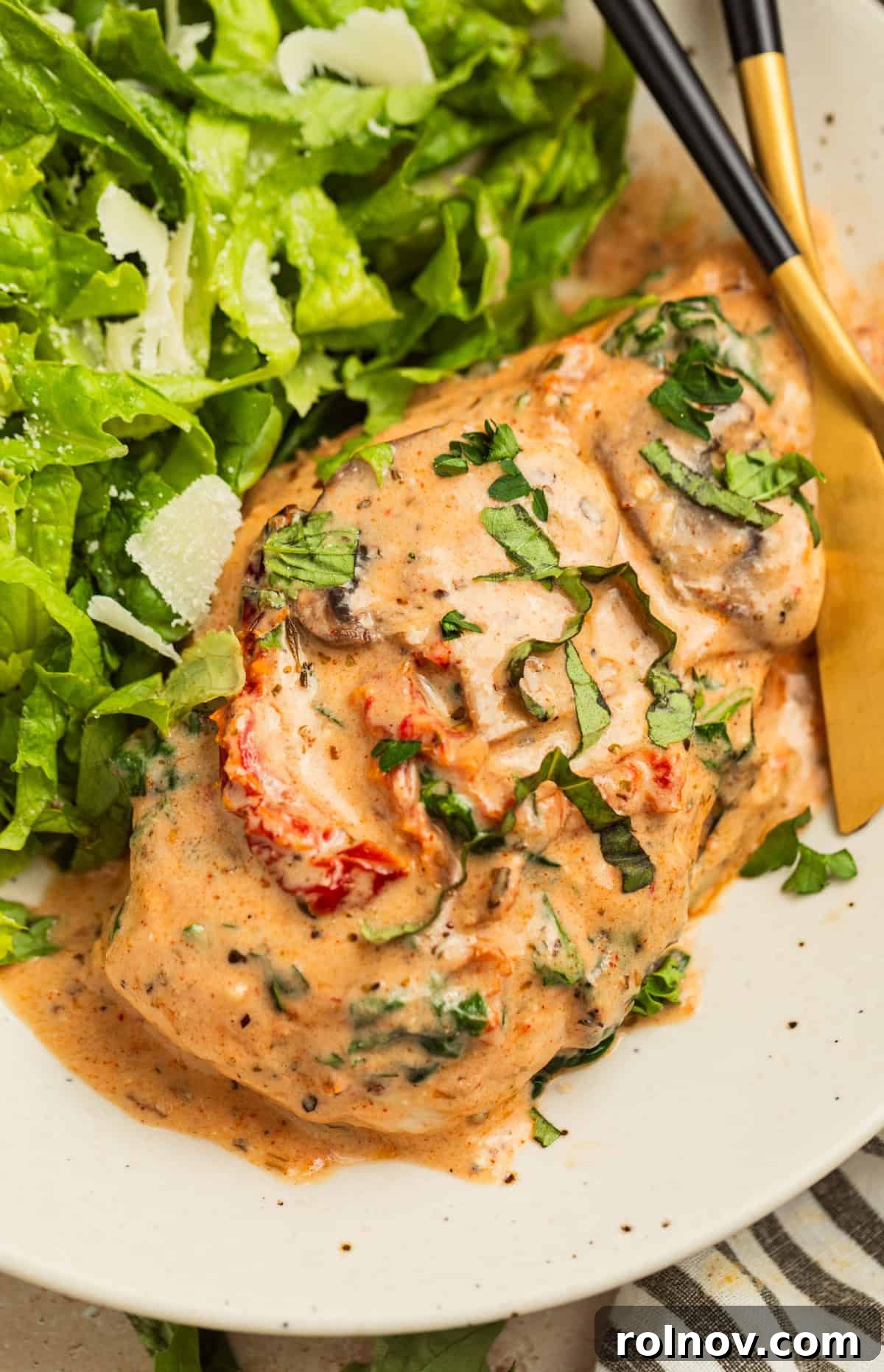 Savory Tuscan Chicken Bake 12 A single portion of creamy baked Tuscan chicken breast on a white plate, garnished with fresh herbs, alongside a fresh green salad with a fork.
