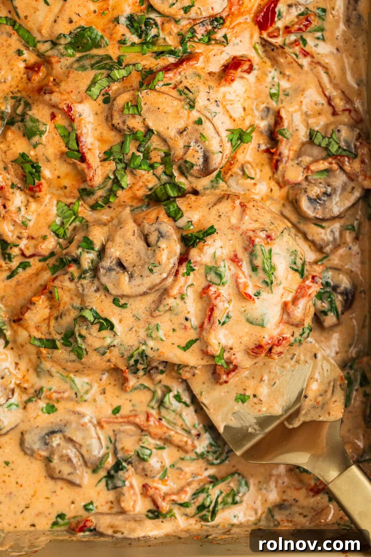 Savory Tuscan Chicken Bake 3 Close-up of baked Tuscan chicken in a pan, smothered in a creamy sauce with visible mushrooms, vibrant green spinach, sun-dried tomatoes, and a sprinkle of fresh parsley.